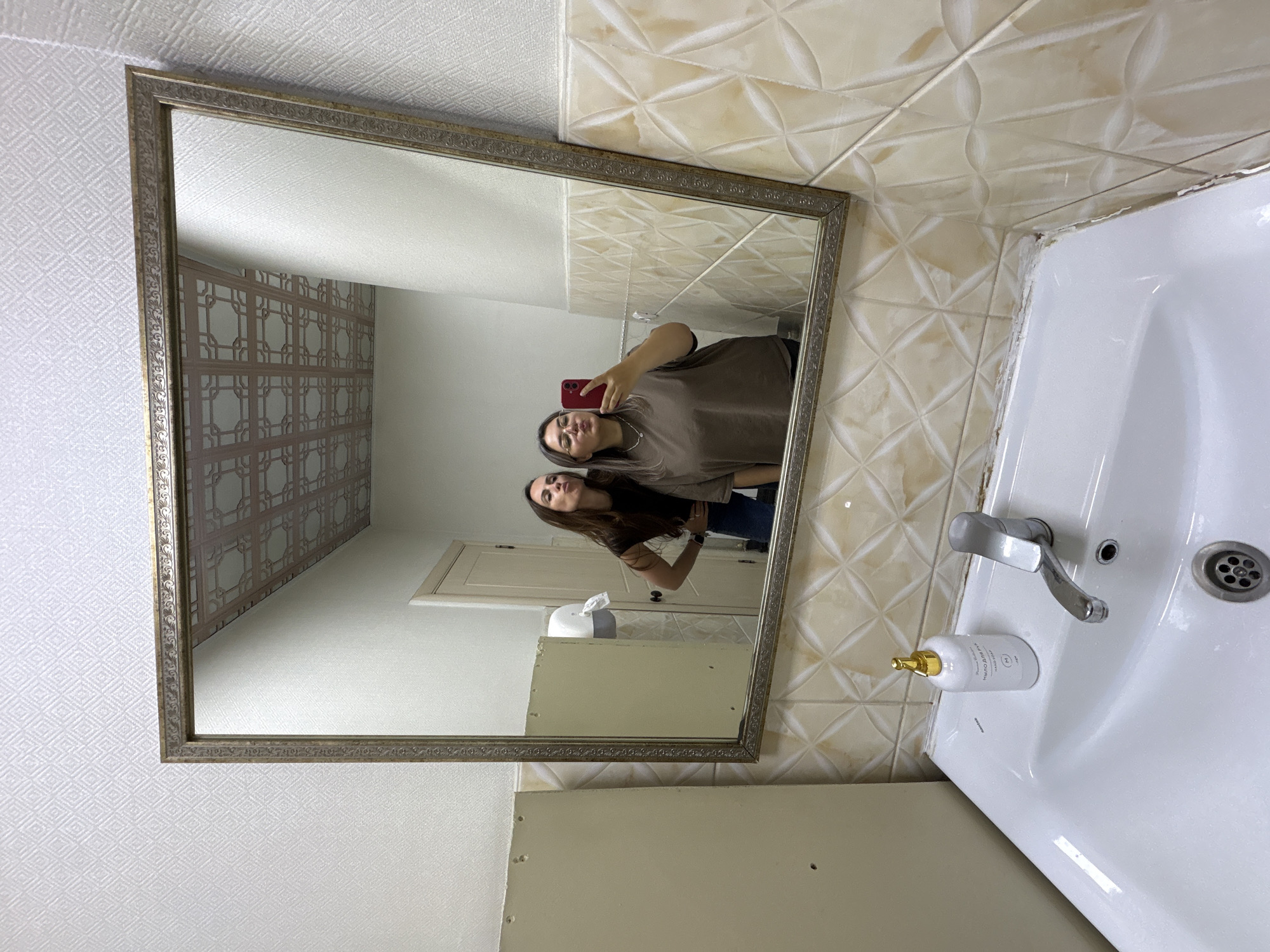 Two women are reflected in a framed mirror in a bathroom. The woman closest to the mirror holds a red phone, seemingly taking a selfie. The other woman stands behind her, with her arm around her. The bathroom features patterned wallpaper, tiled walls, a white sink, a dispenser bottle, and a faucet. The location is unknown.