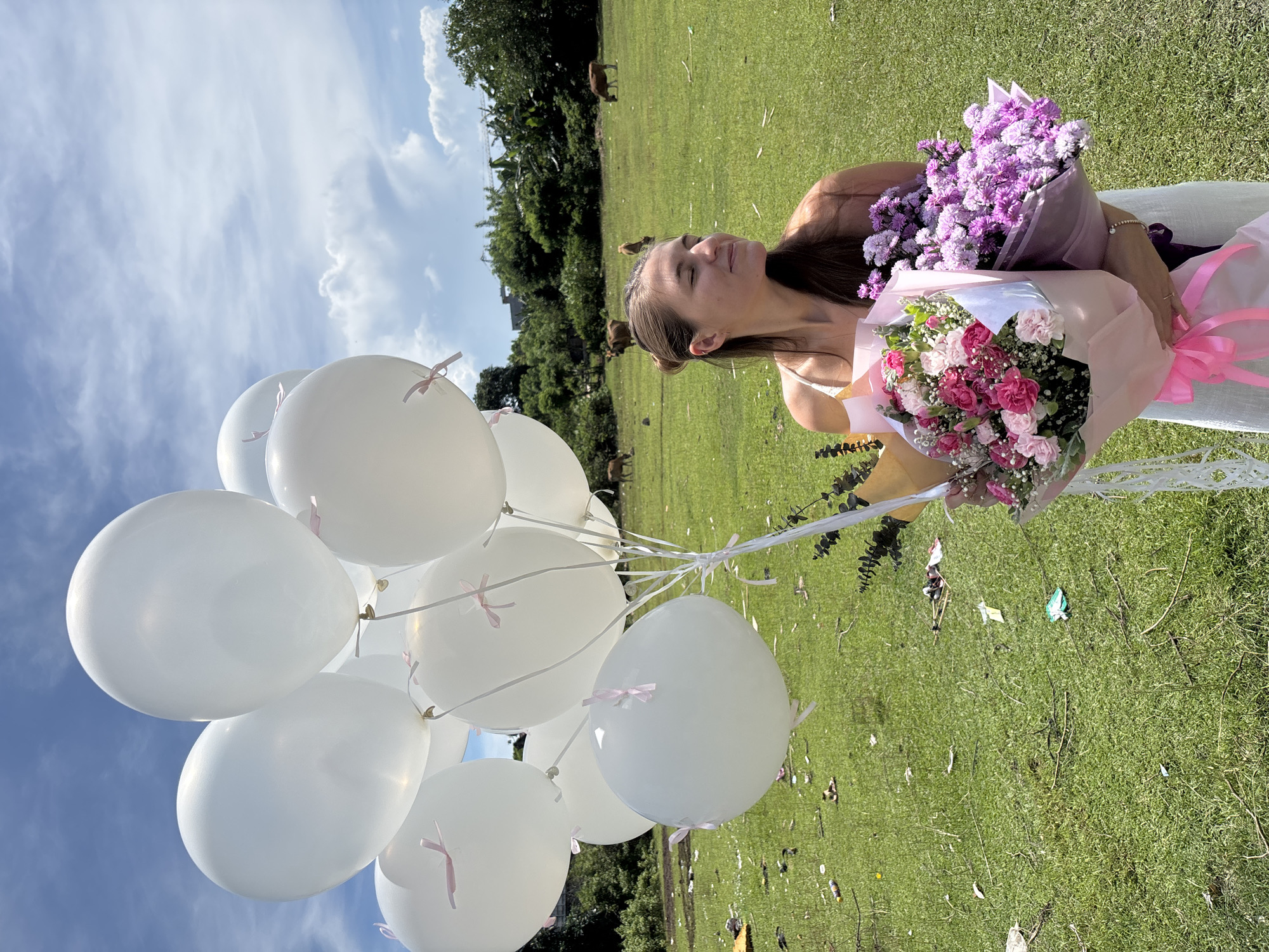 A person stands in a grassy field holding two bouquets of flowers and a bunch of white balloons. The person has light skin, brown hair pulled back in a bun, and wears a light-colored garment. The bouquets consist of pink and red carnations and purple asters. The balloons are tied with pink ribbons. The location is Kuta Selatan, Indonesia. The background shows a field with sparse trees and grazing cows under a sky with some clouds. Debris litters the grass.