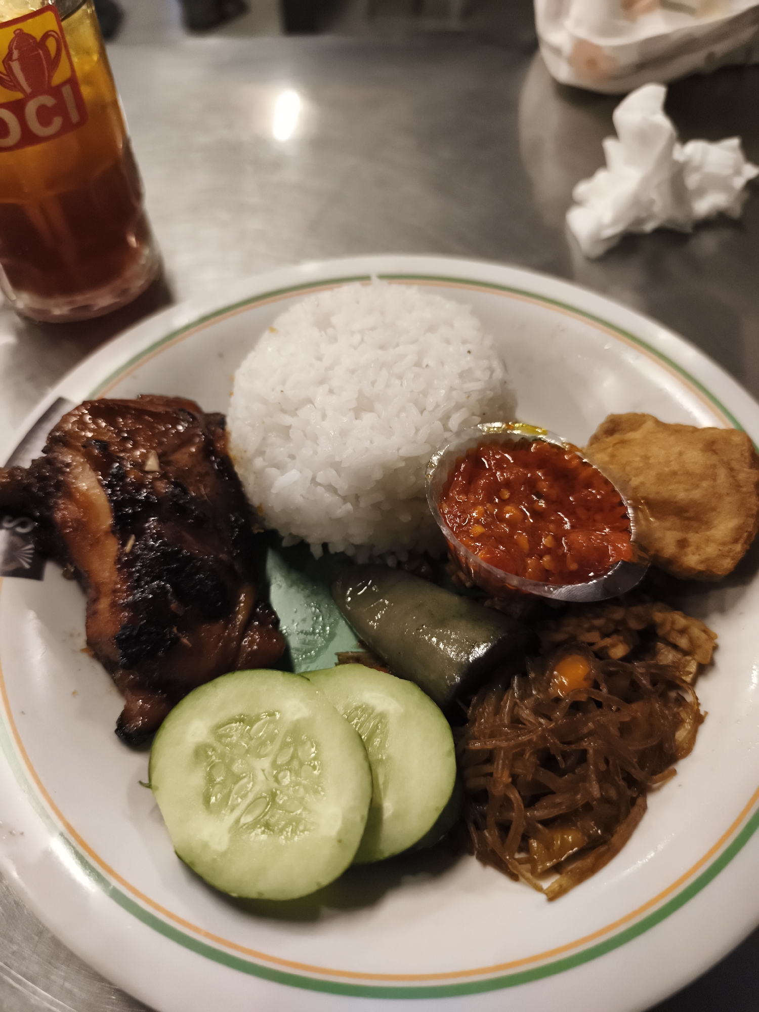 A close-up, eye-level shot captures a plate of Indonesian food, likely served at a restaurant in Sokanegara, Indonesia. The meal consists of white rice, a piece of dark, grilled chicken, a small container of red chili paste, a piece of fried tofu, slices of cucumber, a piece of cooked eggplant, and a side dish of shredded vegetables with orange accents. To the left of the plate, a glass of iced tea with a branded label is visible. The setting appears to be a table with a metallic surface, and a crumpled white napkin is present in the background. The lighting is somewhat dim, casting reflections on the table.