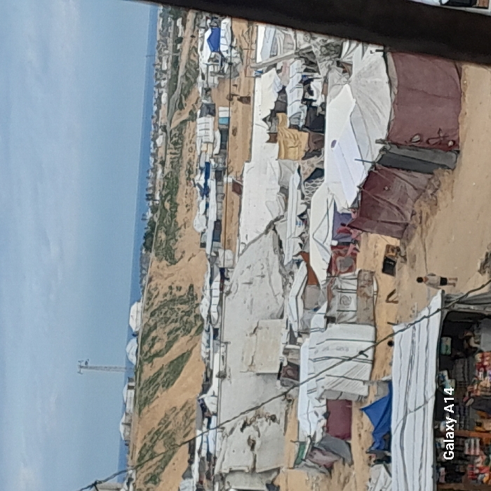 A landscape features an encampment near a coast under a cloudy sky. The settlement is made of tents and makeshift shelters, densely packed on sandy ground. Clotheslines stretch between the structures, and an individual walks through the camp. A tower stands in the distance. The image suggests a temporary or informal settlement, possibly a refugee camp or a community in crisis.