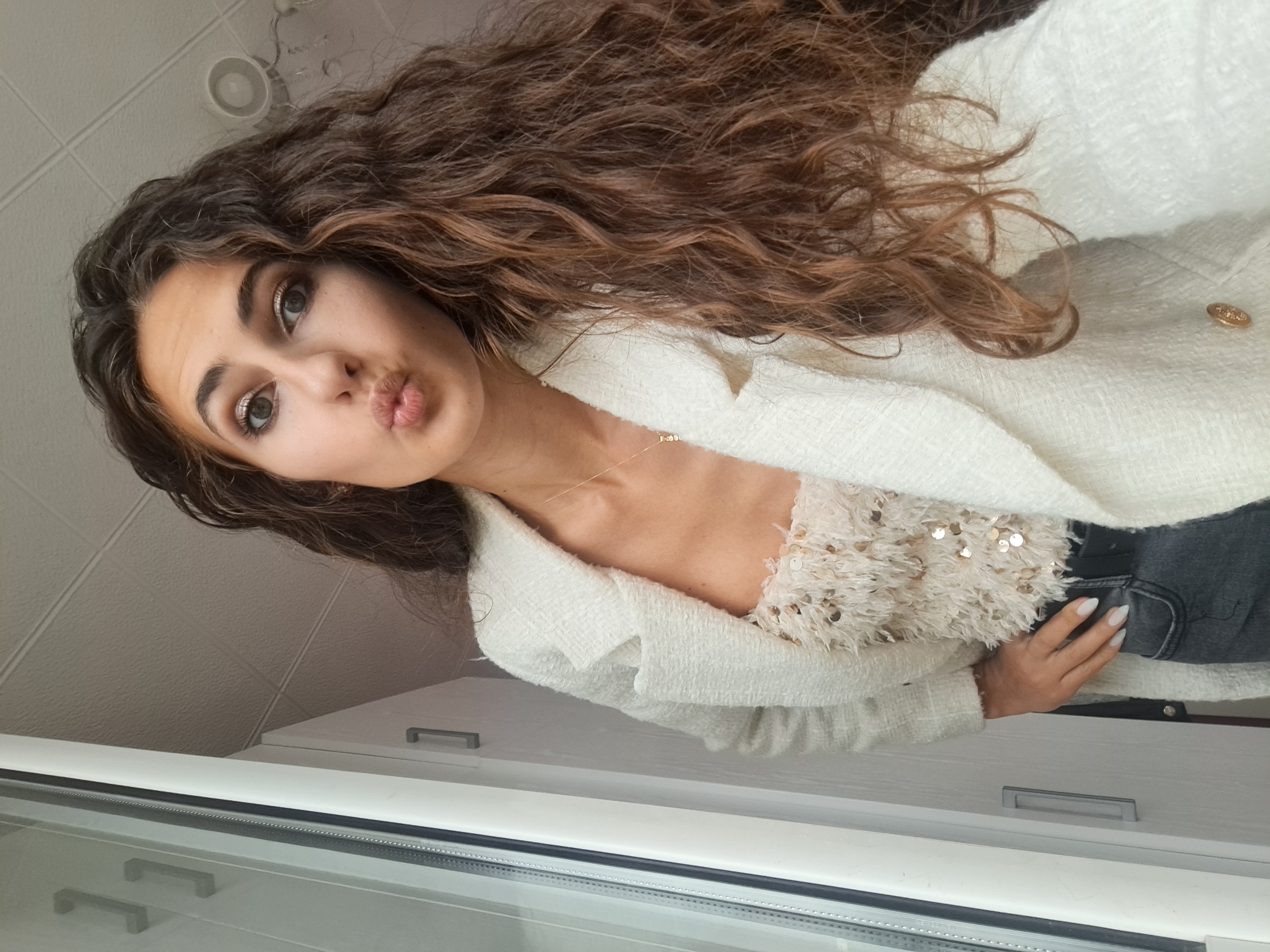 A young woman with long, curly brown hair and a light complexion poses for a selfie indoors. She is wearing a white tweed blazer over a cream-colored, sequined top, and dark grey jeans. Her lips are puckered, and she has makeup on her eyes and lips. She is standing near a window, with a portion of the window frame visible on the left side of the image. The background shows a textured ceiling and a light fixture. There are no indications of the location being in Negovan, Bulgaria.
