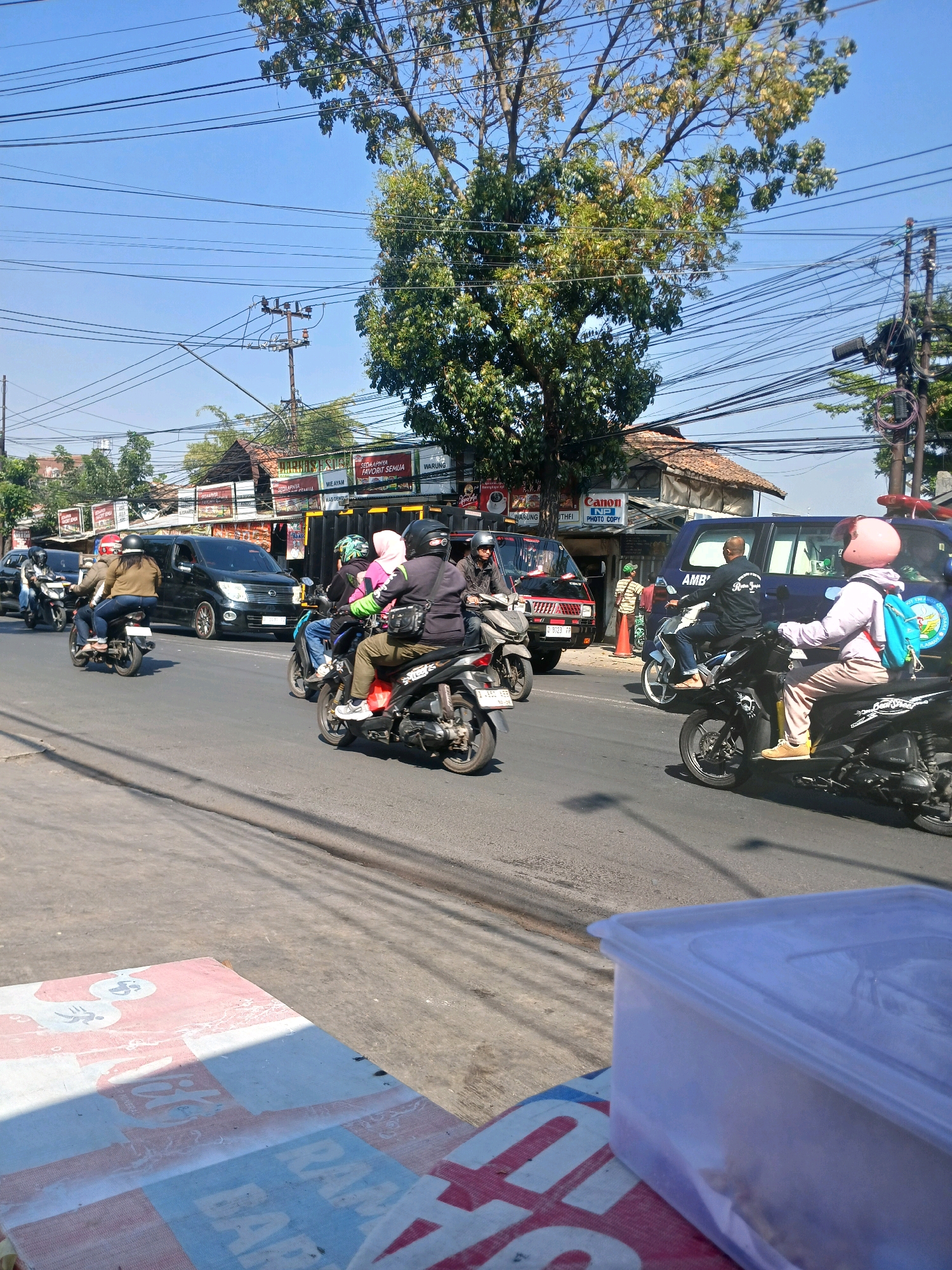 This is a medium shot, eye-level, outdoor, daytime, slightly blurry, vertical photograph of a street scene in Bandung City, Indonesia. The foreground features a plastic container and a red and white patterned cloth on a table, with shadows cast across the surface from the right. The midground displays a road with several motorcycles and a black van. On the motorcycles are multiple individuals wearing helmets and casual clothing, some carrying bags. A person in a light purple jacket and pink helmet is on the rightmost motorcycle, facing away from the camera, with a blue backpack. The background reveals buildings with orange-tiled roofs, trees with green and yellow leaves, and a clear blue sky filled with a dense network of overhead power lines and utility poles. Several signs are visible on the buildings, including "Canon NP PHOTO COPY" and what appears to be a food stall sign. The overall impression is a busy urban street with significant electrical infrastructure.