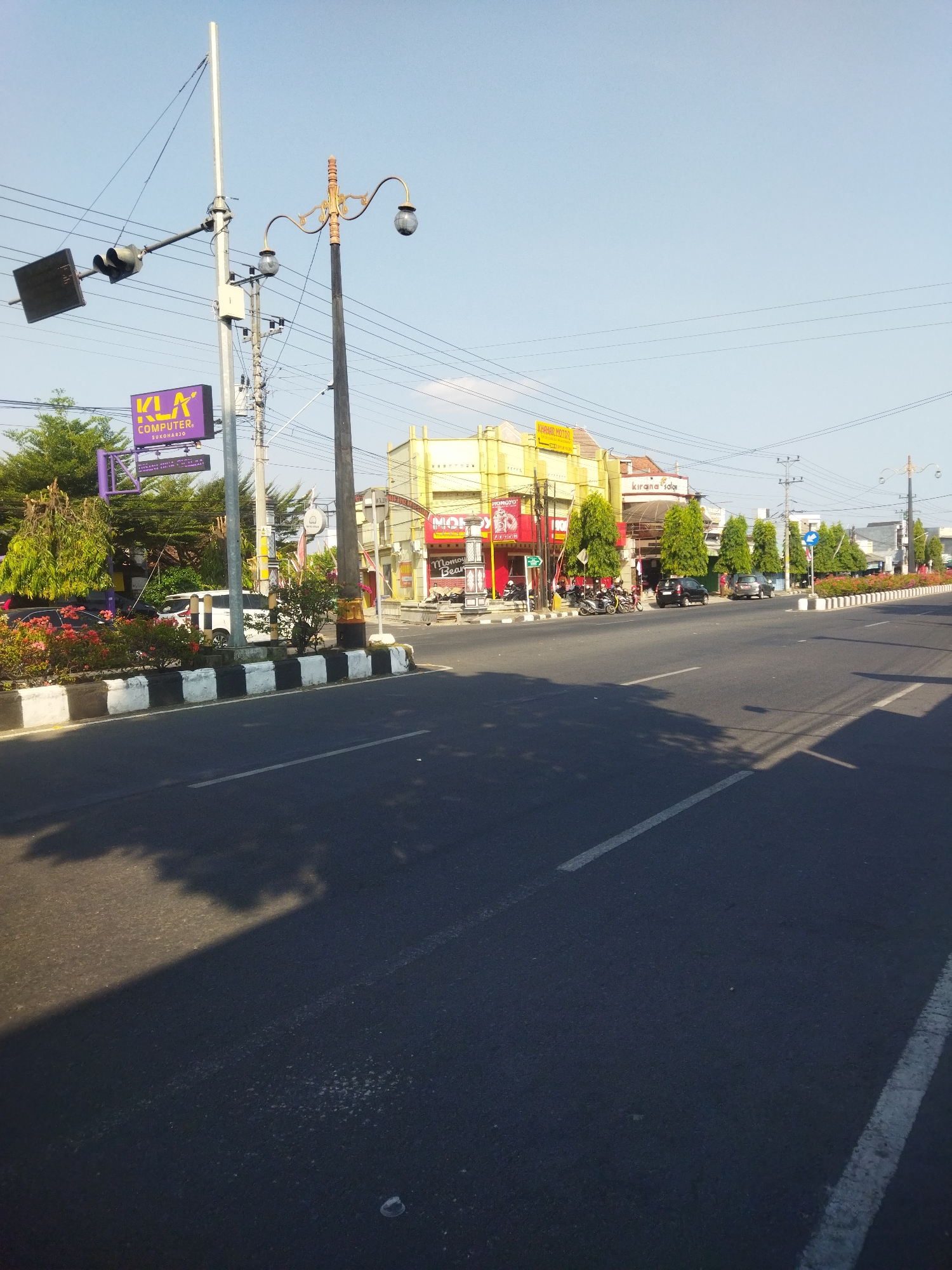 The image depicts a street scene in Sukoharjo, Indonesia. On the left side of the road, there is a traffic light and a tall pole with various electrical wires. A purple sign for "KLA COMPUTER SUKOHARJO" is visible below the traffic light. A median strip separates the road, adorned with black and white striped barriers and blooming flowers. Several cars and motorcycles are parked along the side of the road, and some people are standing near the buildings. The buildings lining the street are colorful, with prominent signage, including a yellow building with a sign that reads "KHADIRI MOTOR" and a red building with "MOMOYA" signage. A street lamp with an ornate design stands on the right side of the road, casting shadows from the trees onto the asphalt. The sky is clear and blue, suggesting it is daytime. The road itself is paved with asphalt and marked with white lines.