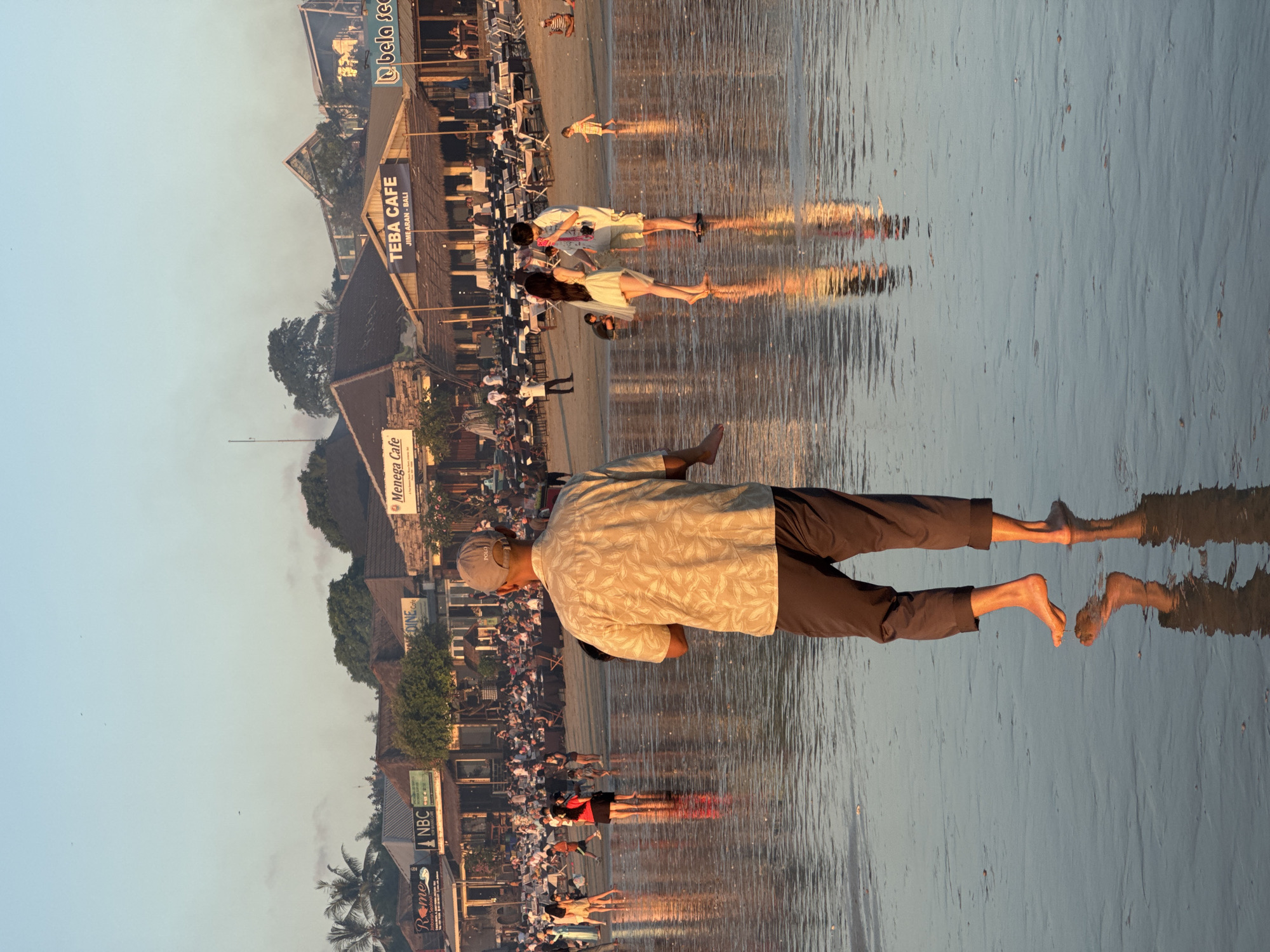 The image depicts a busy beach scene at sunset in Kuta Selatan, Indonesia. In the foreground, a man wearing a patterned short-sleeved shirt, dark pants, and a baseball cap stands with his back to the camera, facing the water. His reflection is clearly visible on the wet sand. Further back, a beach lined with numerous restaurants and cafes is bustling with activity. Several couples and groups of people are visible walking along the shore and sitting at tables. Notable signs include "Menega Cafe" and "TEBA CAFE JIMBARAN - BALI". The sky is clear with a soft gradient of colors, indicating the late hour of the day. The overall atmosphere suggests a vibrant and popular tourist destination.