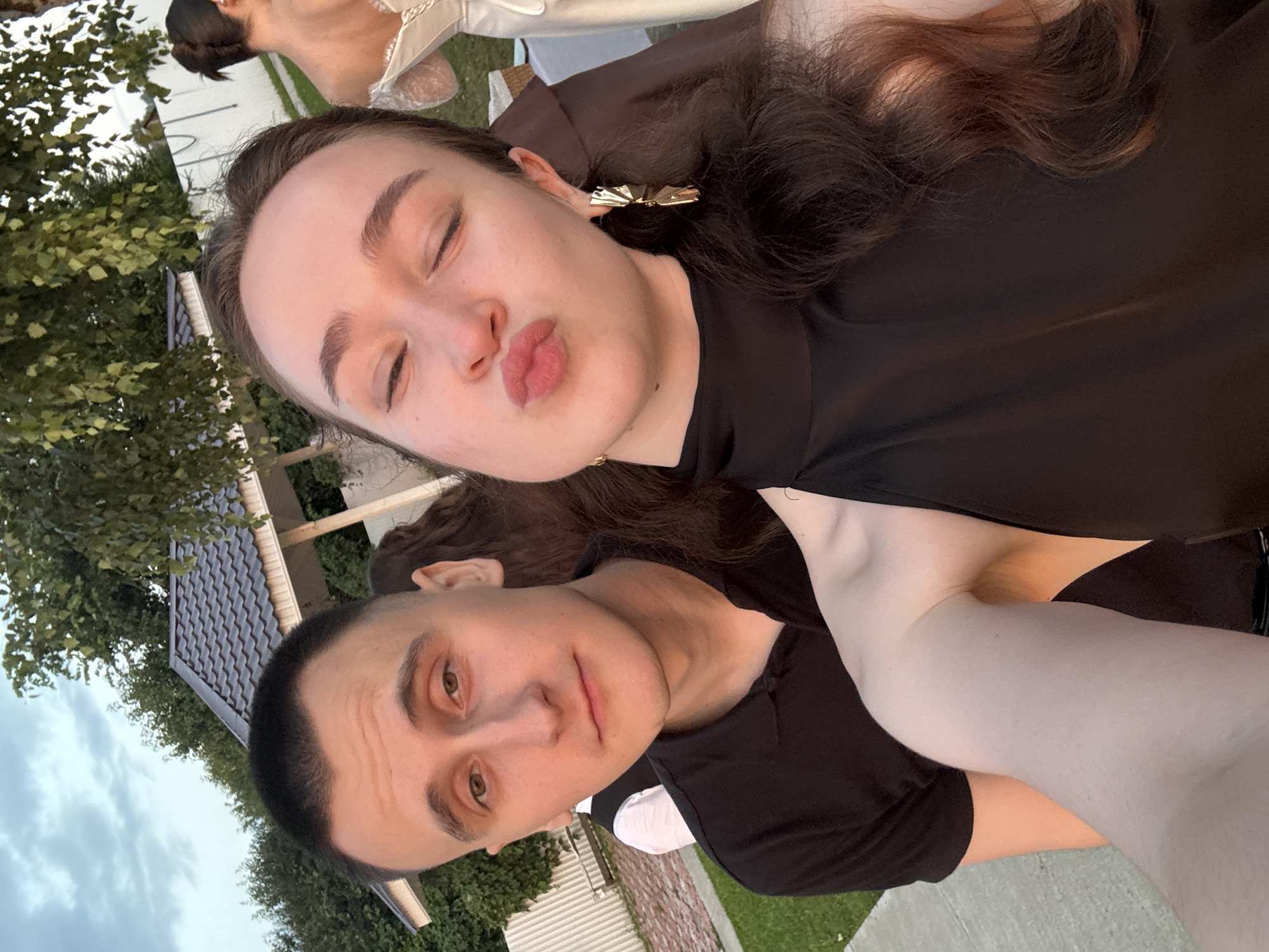This is a selfie of a young couple outdoors. The man on the left is wearing a black polo shirt. The woman on the right is wearing a black dress with a halter top and large gold earrings. She is puckering her lips for the camera. In the background, a woman in a white dress is visible, possibly a bride. Behind them, there is a gazebo with a dark roof, trees, a white fence, and a grassy area. The sky appears overcast. The location is implied to be in the city of Olkhovka, Russia, suggesting a suburban or rural setting rather than a dense urban environment.