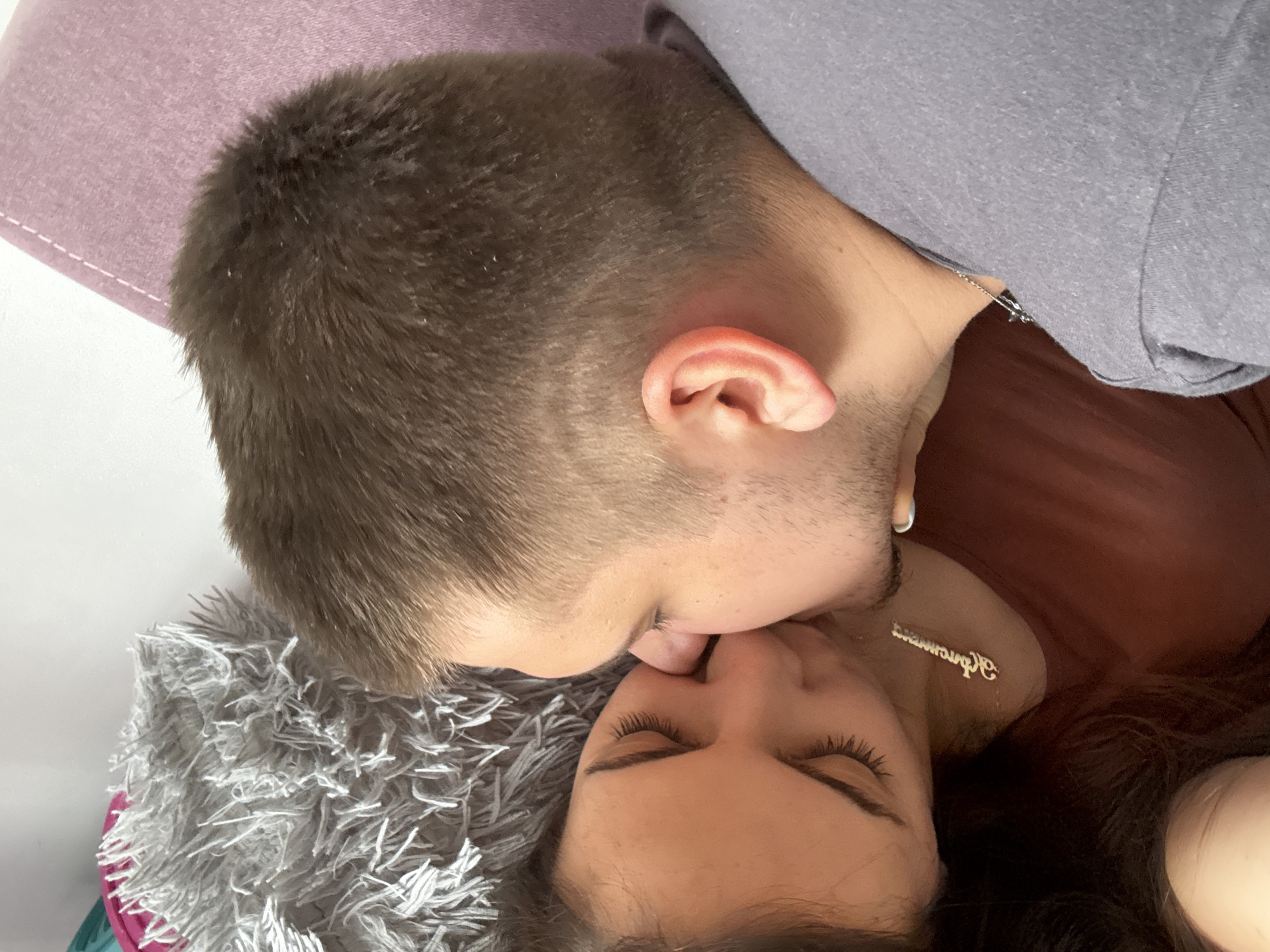 A man and a woman are kissing. The man has short brown hair and is wearing a gray t-shirt. The woman is wearing a brown shirt and a necklace with a word or name on it. They are lying on a gray shag rug or blanket. In the upper left corner is a purple pillow. The location is not identifiable as Plovdiv, Bulgaria based on this image.