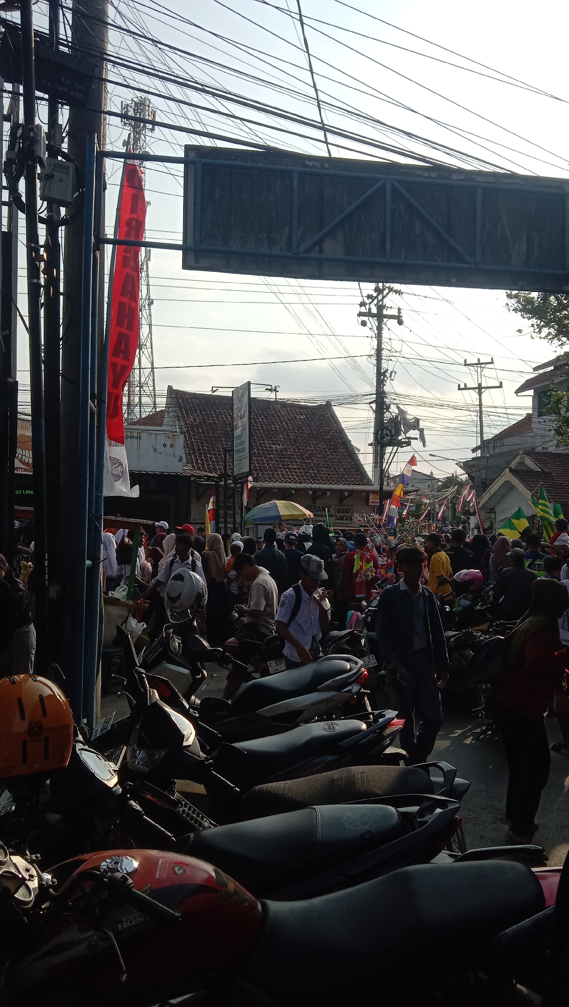 This is a street-level, eye-level, vertical, full-shot photograph taken in the city of Pesantren, Indonesia. The scene depicts a busy street with a crowd of people, parked motorcycles, and various buildings. A large red banner hangs vertically on a utility pole on the left side of the image, with Indonesian text visible. In the background, several buildings with red tile roofs are present, along with numerous electrical wires and poles. People are walking and gathered in groups, many wearing head coverings. Several motorcycles are parked in rows in the foreground and middle ground. The sky is visible at the top of the image, appearing partly cloudy. The overall impression is of a public gathering or event taking place in a densely populated urban area.