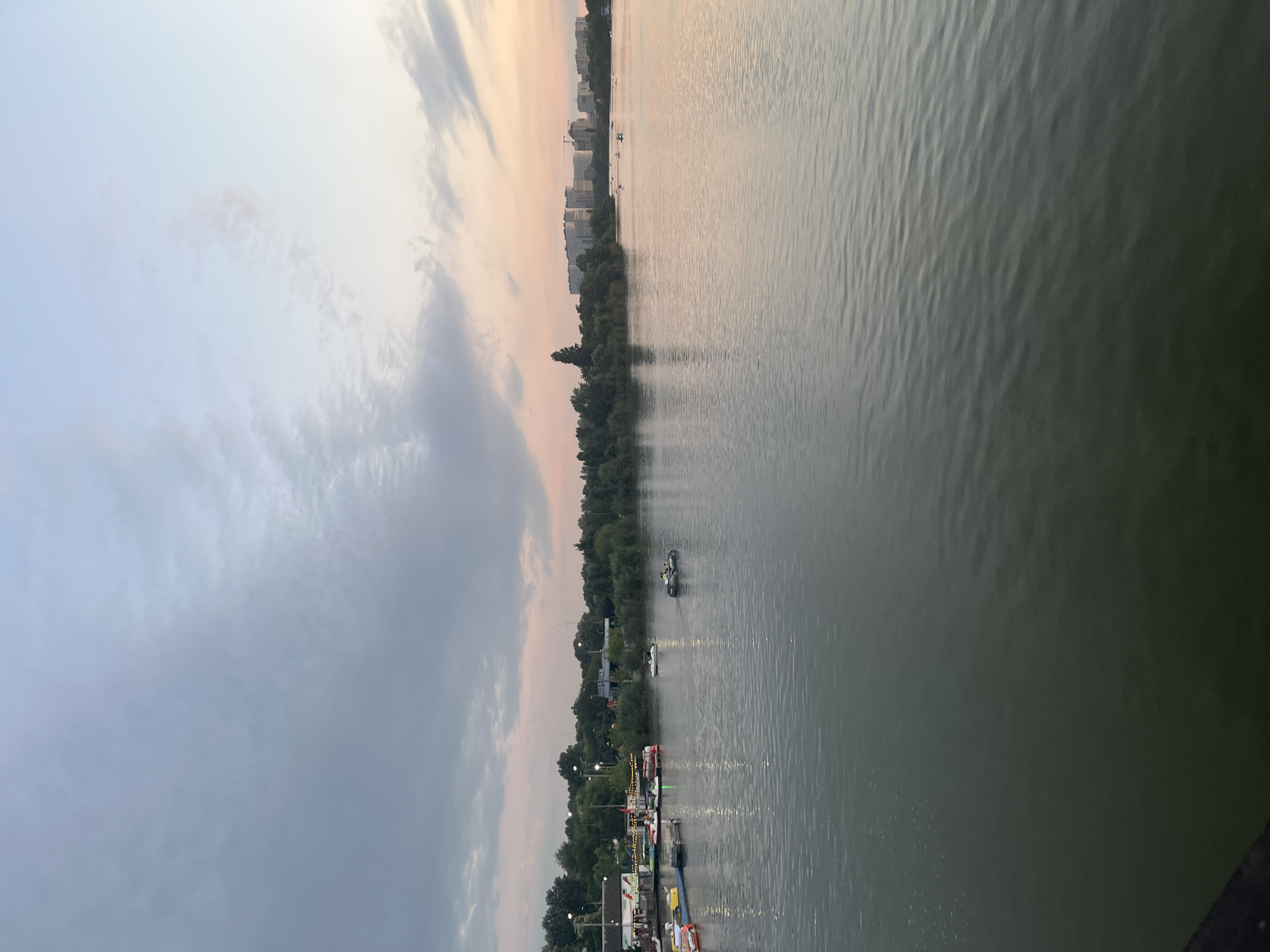 This image depicts a view of the Kuban River in Krasnodar, Russia, during a twilight hour. The sky displays a gradient of colors, transitioning from pale blue and grey clouds to soft pink and orange hues near the horizon, indicating either sunrise or sunset. On the left side of the riverbank, there is a stretch of green trees and a few buildings, including what appears to be a pier or a waterfront recreational area with several small boats docked. Some of these boats are pedal boats. Further out on the water, there are a few individuals in boats, some of which appear to be pedal boats, and others might be small motorboats or kayaks. In the background, across the river, a line of modern residential buildings and construction cranes are visible, indicating an urbanized area. The water's surface is calm with gentle ripples.