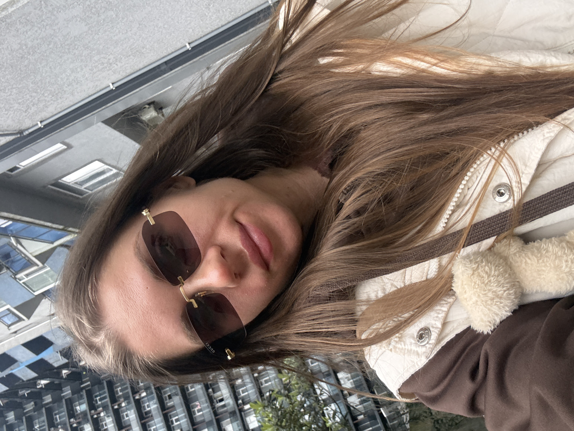 A young woman is posing for a selfie outdoors in Batumi, Georgia. She is wearing rimless sunglasses with dark lenses and gold frames. The glasses have "GUCCI" on the side. She has long, brown hair that is partially covering her face. She is wearing a brown sweatshirt with a beige and brown lined jacket. The jacket has fur trim. She is outdoors in an urban environment, with a tall building visible behind her and greenery behind that. The buildings are of a modern style and appear to be a high-density residential or commercial area. The image suggests a cool, sunny day in Batumi.