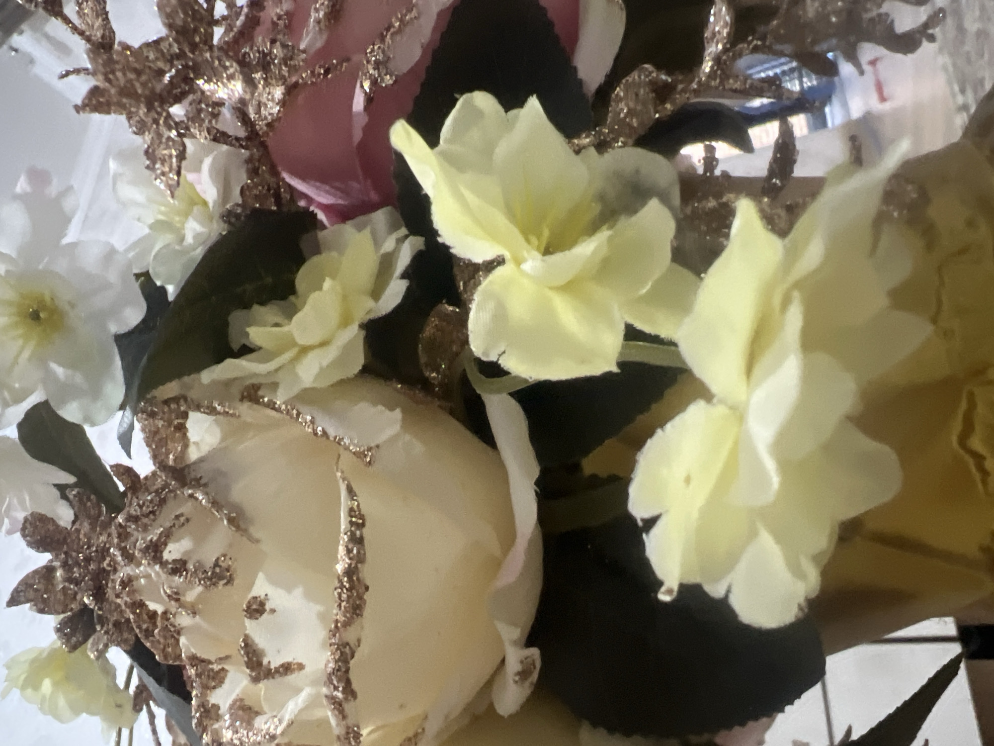 A close-up, eye-level, medium shot captures a floral arrangement. The arrangement features several artificial flowers, predominantly in cream and pale yellow hues, with one pink flower partially visible on the right. The petals of the cream-colored roses are edged with gold glitter. Several small, delicate, pale yellow flowers are also present, some with multiple layers of petals. Dark green leaves are interspersed throughout the bouquet, providing a contrast to the lighter-colored blooms. Decorative gold glittery sprigs are woven among the flowers, adding a festive touch. The flowers are displayed in a gold-colored vase, which is visible at the bottom of the frame. The background is blurred, suggesting an indoor setting, possibly a home or event space, with hints of furniture and lighting.
