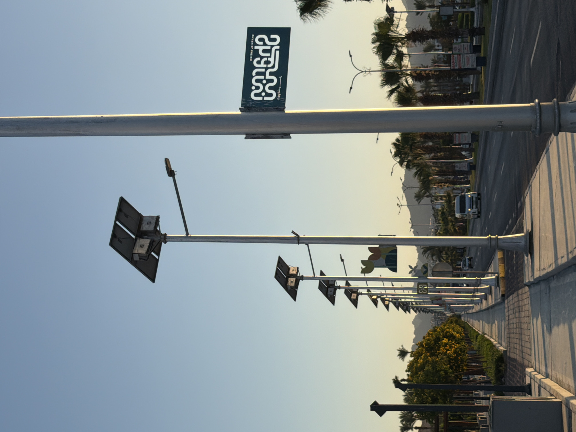 The image shows a street scene in Sharm El Sheikh, Egypt, with a clear blue sky. The foreground features a sidewalk and a road. Along the sidewalk, there are multiple lampposts equipped with solar panels. One lamppost has a sign with Arabic text and the words "SHARM EL SHEIKH" printed horizontally. The road has a few vehicles, including a white van. Palm trees and lush green foliage line the street, with mountains visible in the background. The sun appears to be low in the sky, casting long shadows.