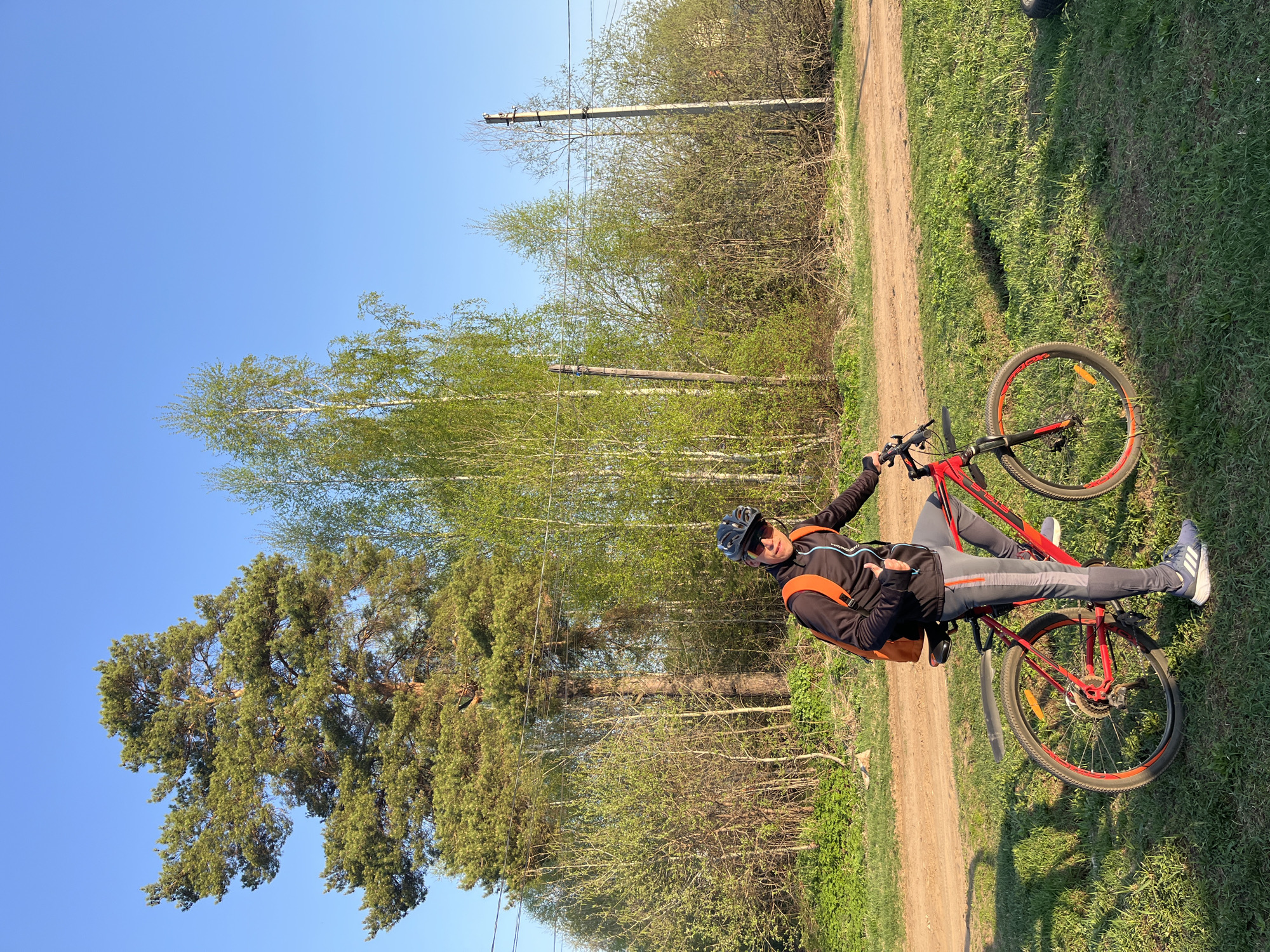 A person rests on a red mountain bike along a dirt path, likely in a park. The person wears a dark jacket with orange accents, gray pants, athletic shoes, a helmet, and sunglasses. The person is reclined back on the bike, facing the camera, with their right hand on the handlebars. The background includes tall trees and clear blue sky. Power lines run above the trees. The location may be in Chaikovsky, Russia.