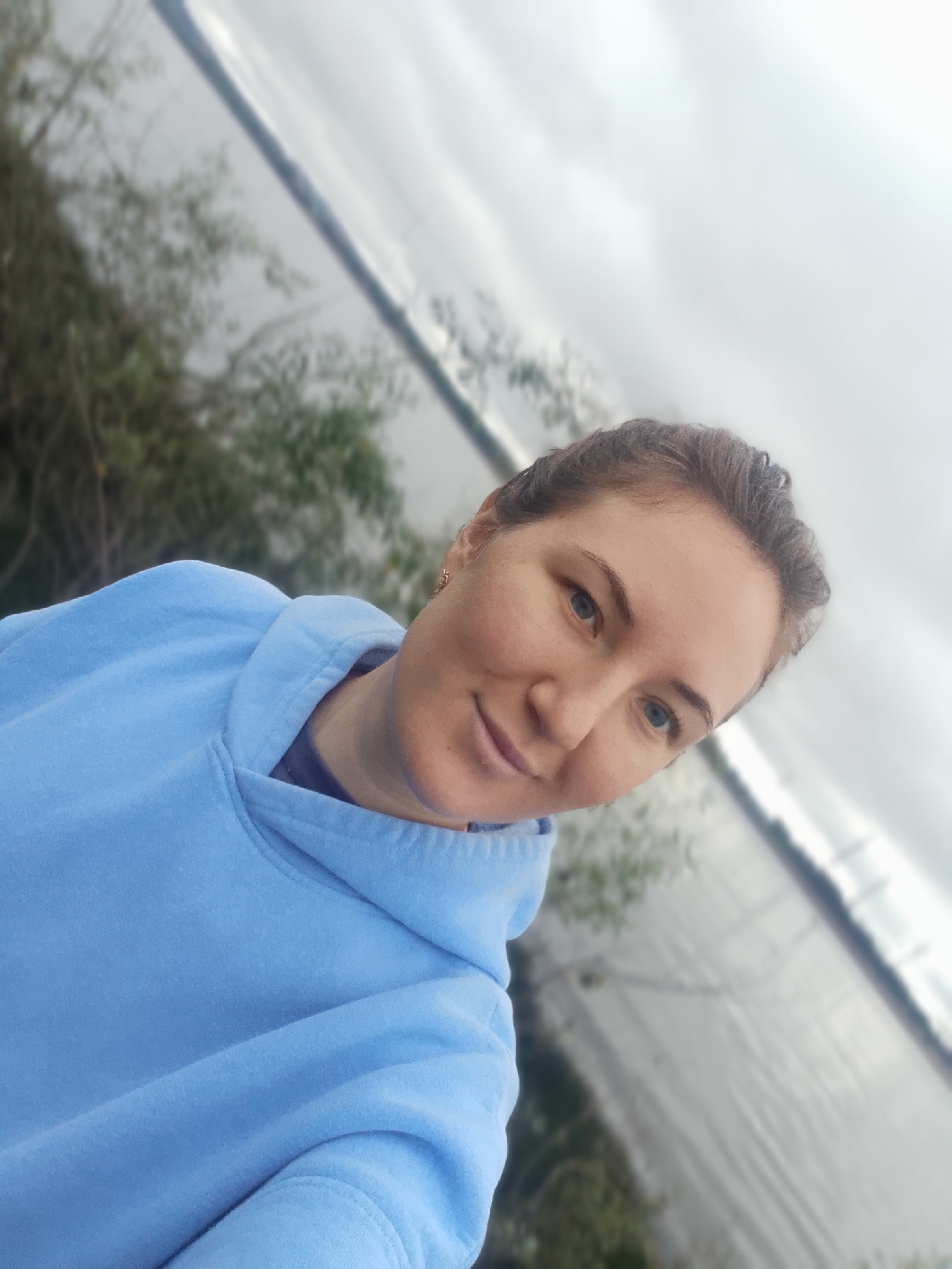 A close-up, slightly tilted selfie captures a young woman with light skin, brown hair pulled back, and blue eyes. She is wearing a light blue hooded sweatshirt. The background is blurred, showing a body of water, possibly a river or lake, with a cloudy sky overhead. Some greenery is visible to the left. The image appears to have been taken outdoors.