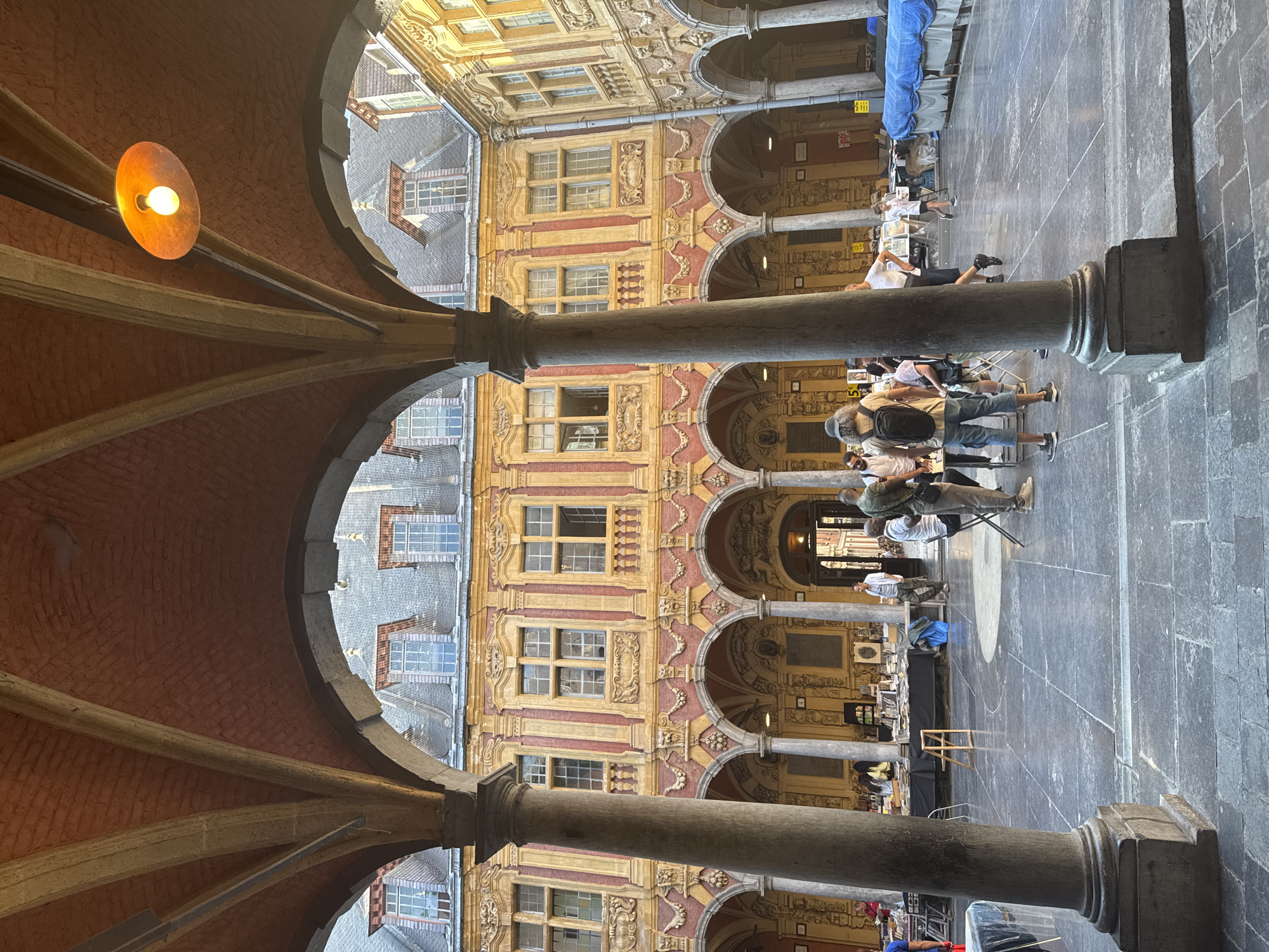 This image depicts an outdoor market or fair held within the courtyard of the Vieille Bourse in Lille, France. The architecture of the surrounding buildings, characterized by arcades, ornate facades with yellow and red detailing, and numerous windows, is clearly visible. Several people are present, browsing stalls set up in the courtyard. Some individuals are seen standing, while others are walking or interacting. The market appears to be selling books and possibly other items, as indicated by the tables laden with merchandise. The ground is paved with large, dark flagstones. An overhead lamp is visible, casting a warm glow.