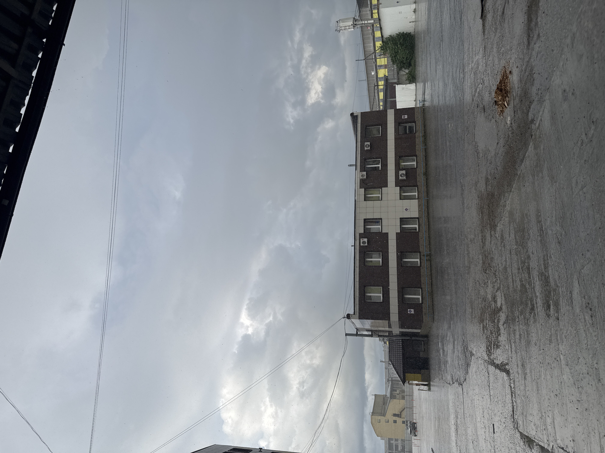 The image displays an urban industrial or commercial area under a heavily overcast sky, with visible precipitation (sleet or light snow/rain) actively falling. The ground, primarily a vast concrete or asphalt expanse, is saturated and reflective due to the moisture, showing numerous cracks and uneven patches. A small mound of dried brown leaves or debris is located on the ground in the mid-right foreground.
The central building is a two-story structure featuring a dark brown lower facade and a lighter beige upper section, horizontally divided by a thin blue band. It has multiple rectangular windows with white frames and several wall-mounted air conditioning units beneath the upper-story windows. A smaller, dark-roofed structure is attached to its left side.
In the background, to the left, stands a light yellow multi-story building with numerous square windows. A dark-colored pickup truck is parked in front of it, near orange barriers. Further in the background, to the right, are additional industrial buildings, some yellow and white, and a tall metal tower with a red light. A small cluster of dark green foliage is visible to the right of the central building. Overhead, multiple power lines traverse the sky. No individuals are present in the scene. The architectural style and industrial landscape are characteristic of an industrial or commercial district in a city like Perm, Russia.