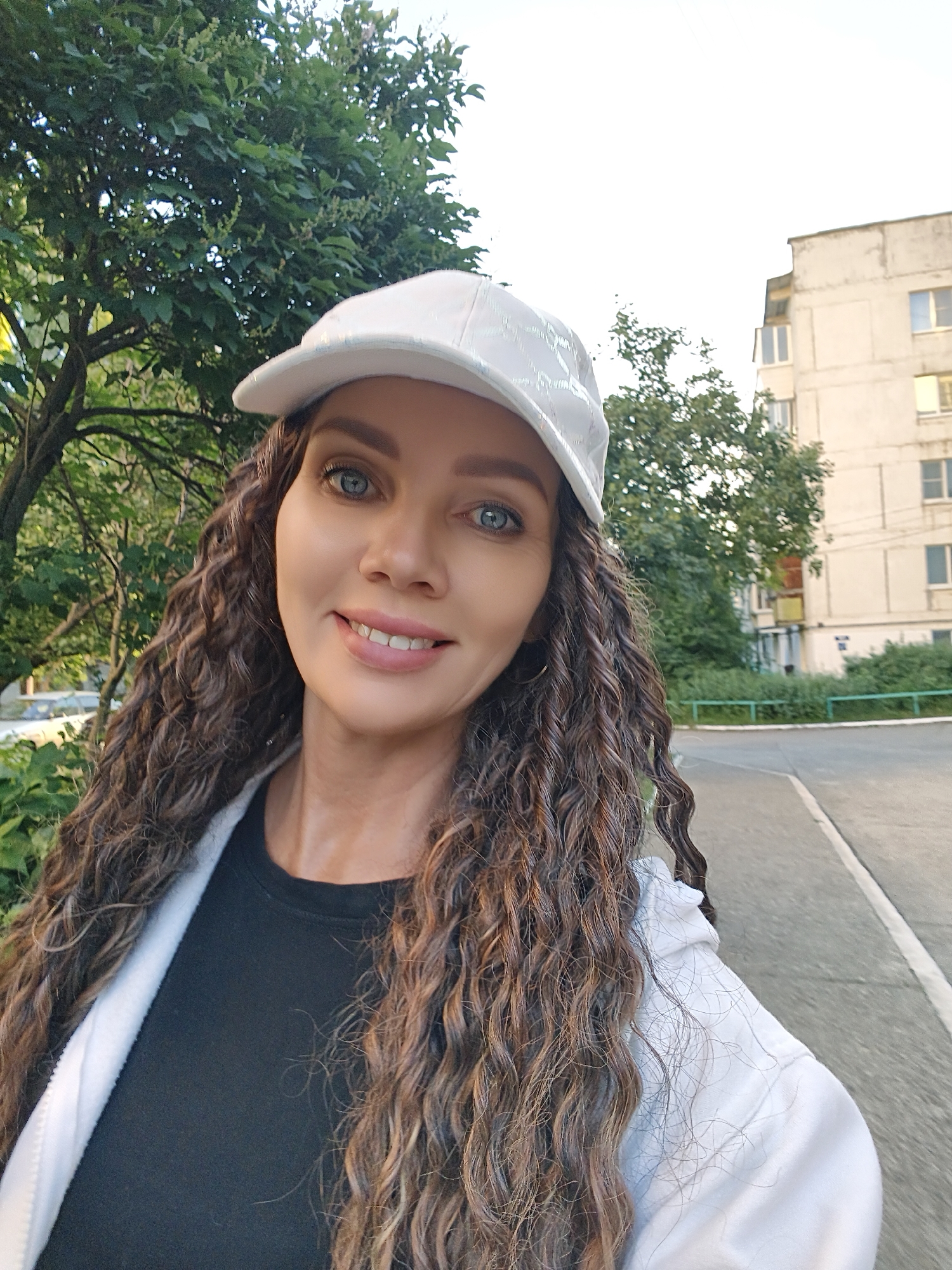 A woman with long, curly brown hair and blue eyes is shown in a selfie. She is wearing a white baseball cap, a black t-shirt, and a white jacket. She is smiling and looking at the camera. The background appears to be an urban setting with trees and a building.