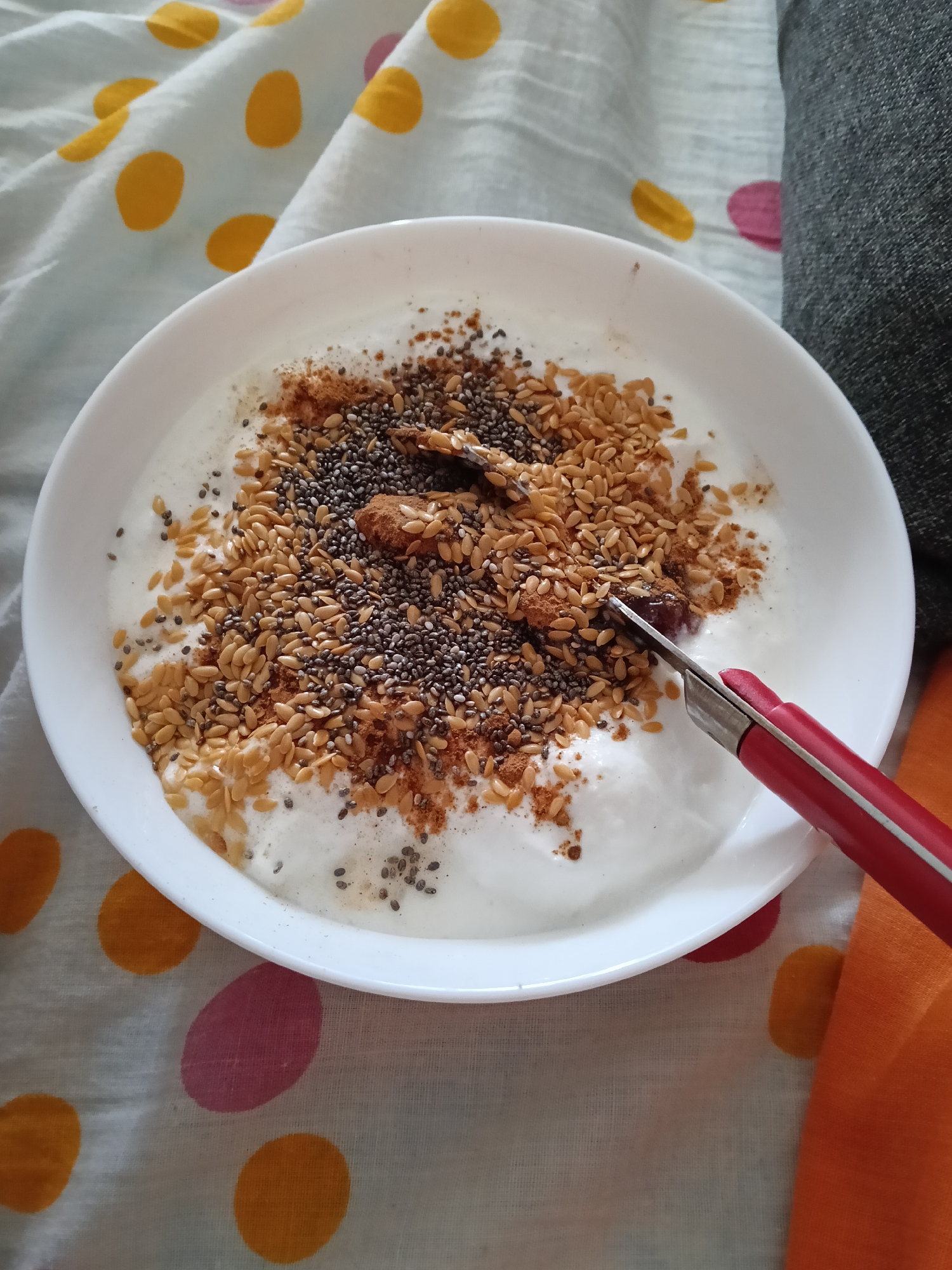 A high-angle, close-up shot shows a white bowl filled with white yogurt topped with a scattering of brown flax seeds, black chia seeds, and reddish-brown cinnamon powder. Embedded in the yogurt are two sliced dates. A pair of small red and silver scissors rests in the bowl, blade-downward, near the right side. The bowl sits on a patterned fabric with yellow and pink polka dots on a white background. The edge of a gray fabric and an orange fabric are visible to the right of the bowl. The location is unknown, and cannot be determined to be San Bartolomé de Tirajana, Spain from the image.
