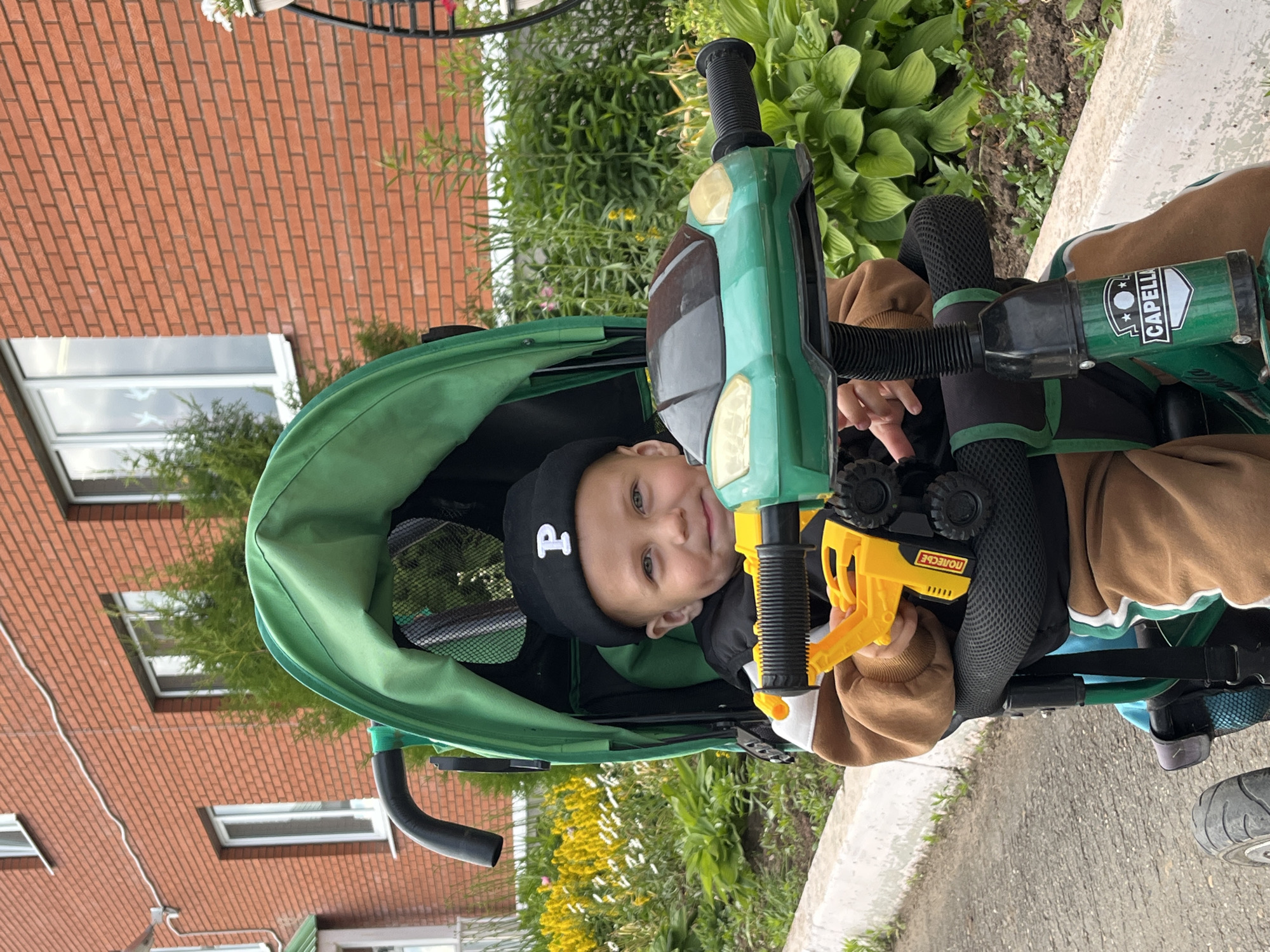A young child is sitting in a green stroller with a black hat on, a yellow toy in their hand, and is looking at the camera. The child is in front of a brick building with windows. The scene appears to be outdoors, possibly in a residential area, with some greenery and a paved surface. The stroller has a green canopy and a green handlebar. The child is wearing a brown hooded jacket and brown pants. The toy in the child's hand is yellow with black wheels. The background shows a brick building and lush green foliage. A portion of a white concrete curb is visible in the foreground.