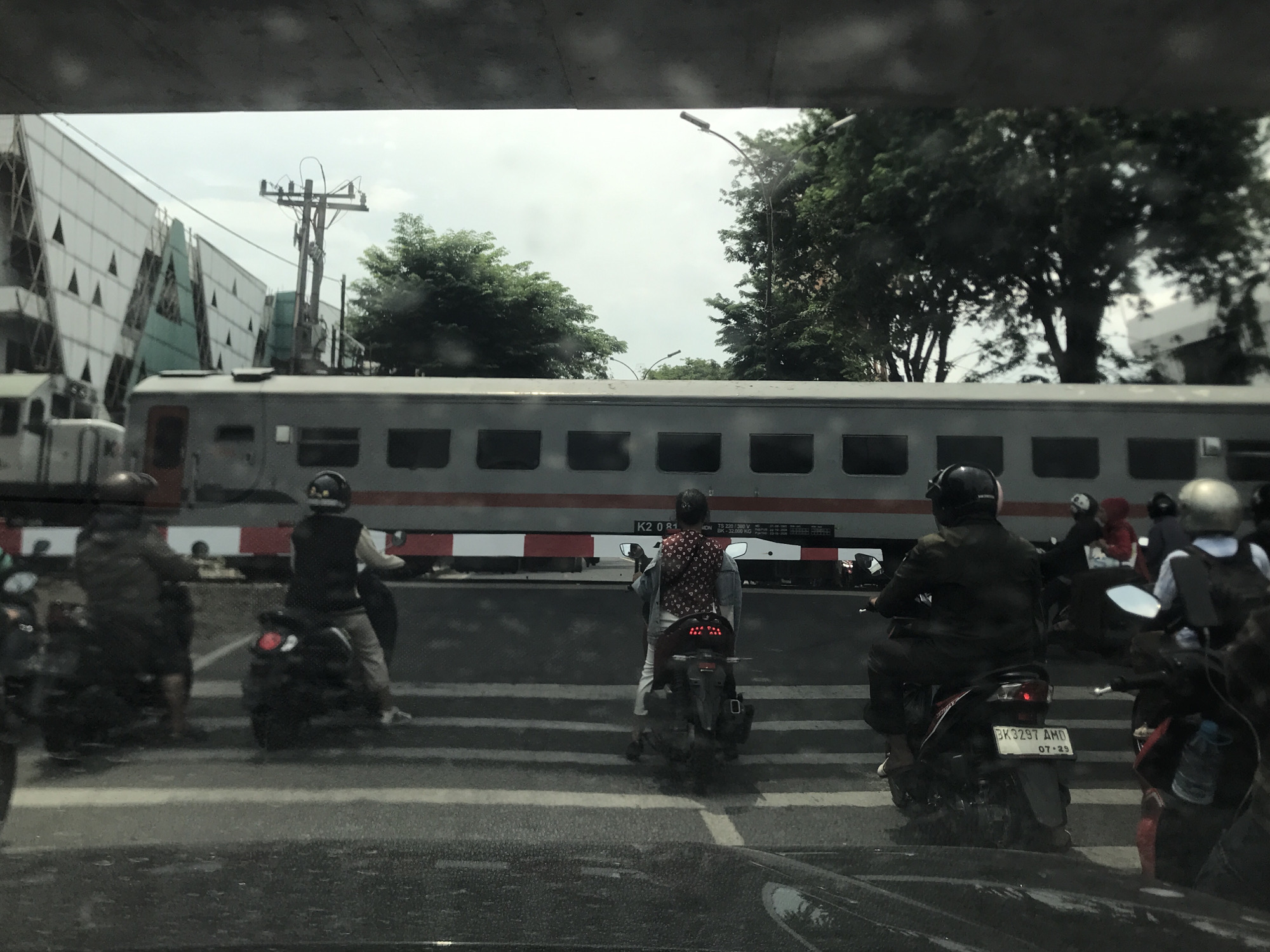 The image, taken in the city of Medan, Indonesia, shows a street scene with several motorbikes stopped at a zebra crossing in front of a passing train. The train is painted grey with white horizontal stripes. The motorbike riders are wearing helmets. A power pole is visible in the background. The photo appears to be taken from inside a car, looking out. A building with a unique triangular design is partially visible. There are several trees present, and the sky appears overcast. The motorbike license plate "BK 3297 AMD" with the date "07-29" is visible.