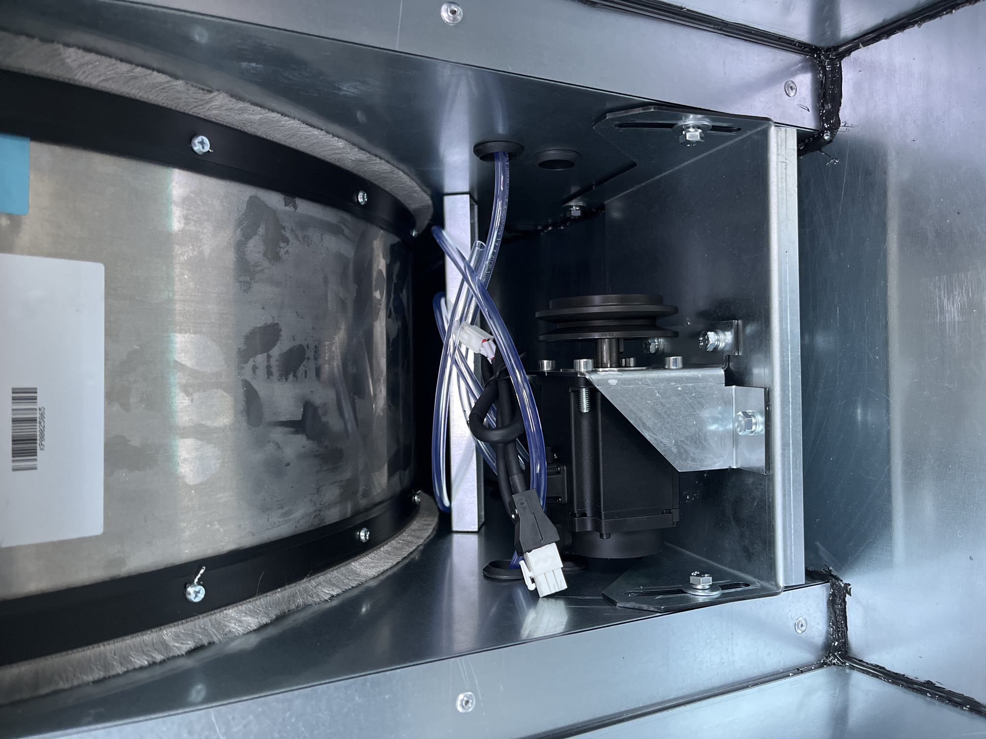 The image shows the interior of a metal enclosure, possibly part of an industrial machine or appliance. A black motor with a pulley attached is visible, mounted on a silver metal bracket. Clear plastic tubing runs from the motor area and passes through a black rubber grommet in the side of the enclosure. The enclosure's interior is lined with a fuzzy, grey material along the top edge. Several screws and bolts are visible, securing different components. The bottom of the enclosure appears to be a flat metal surface. There are no people present in the image. The image does not provide any information about its location in Kyiv, Ukraine.