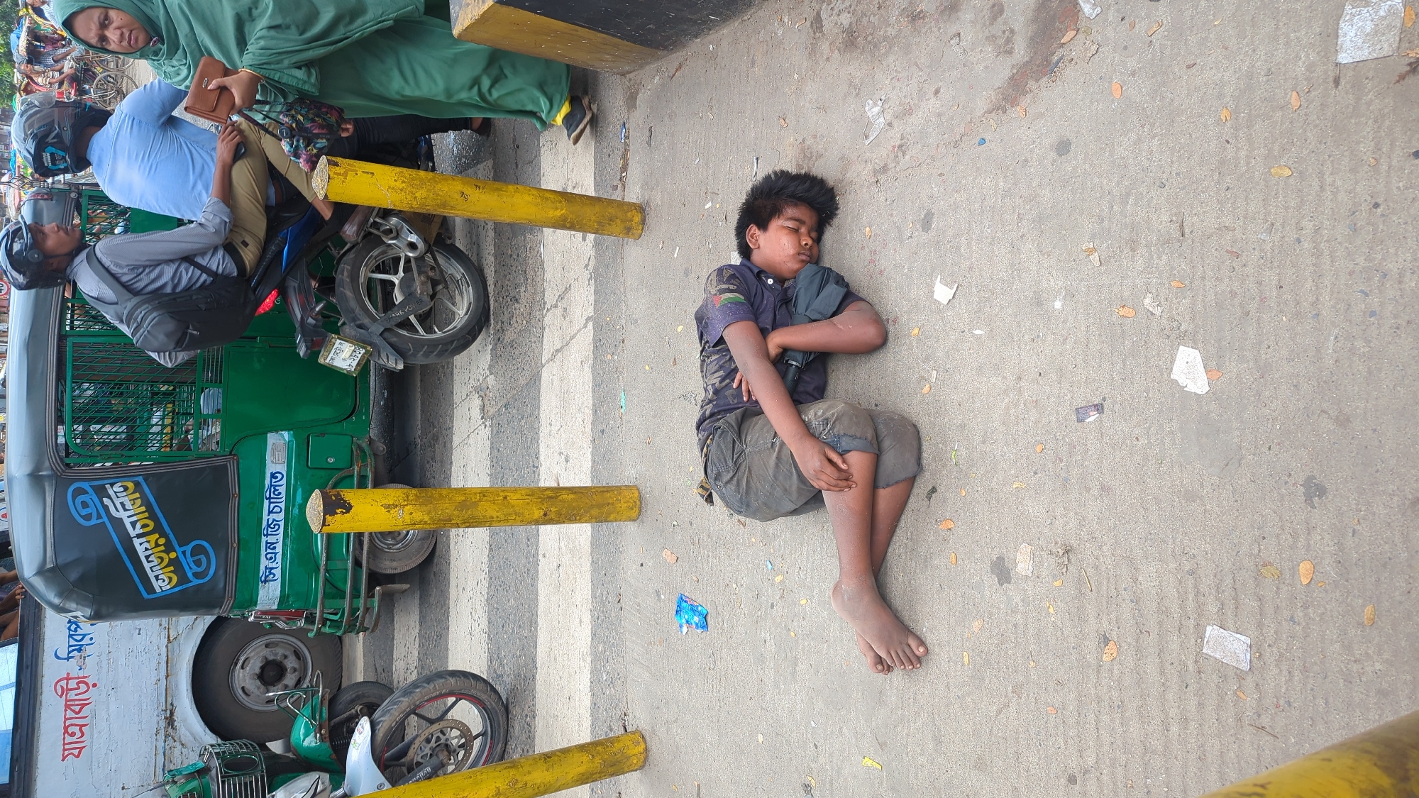 A young boy lies asleep on a sidewalk in Dhaka, Bangladesh, with a street scene unfolding behind him. He is curled on his side, wearing a dark t-shirt and grey shorts, with his face resting on his arm. His feet are bare and dirty. The ground around him is concrete, scattered with litter such as plastic wrappers and fallen leaves.
In the background, a busy street scene is visible. A green auto-rickshaw is stopped, with a bus to its left. Two men on a motorcycle are also present, one wearing a blue shirt and the other a grey shirt. A woman in a green hijab stands to the right, holding a brown wallet. Yellow bollards are positioned on the sidewalk. The overall environment suggests a bustling urban setting, possibly a street intersection or waiting area.