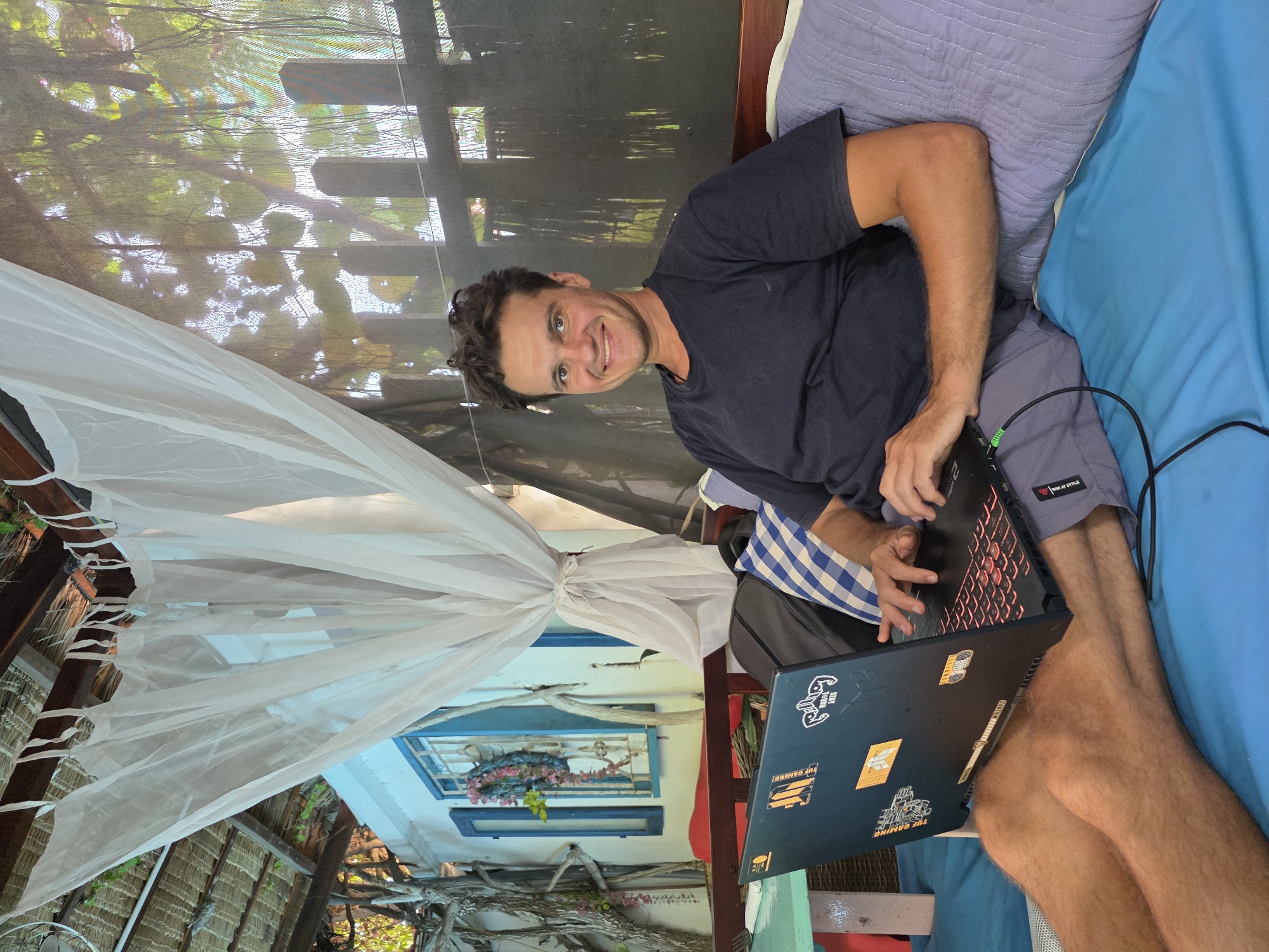 The image shows a man sitting on a blue bed in what appears to be a tropical location. He is working on a laptop, which is resting on his legs. The man is wearing a dark t-shirt and grey shorts. Behind him, there's a window or opening covered by a thin, white curtain. The architecture suggests an outdoor or semi-outdoor setting. The location appears to be in Mũi Né, Vietnam, based on the style of construction and the tropical vegetation.