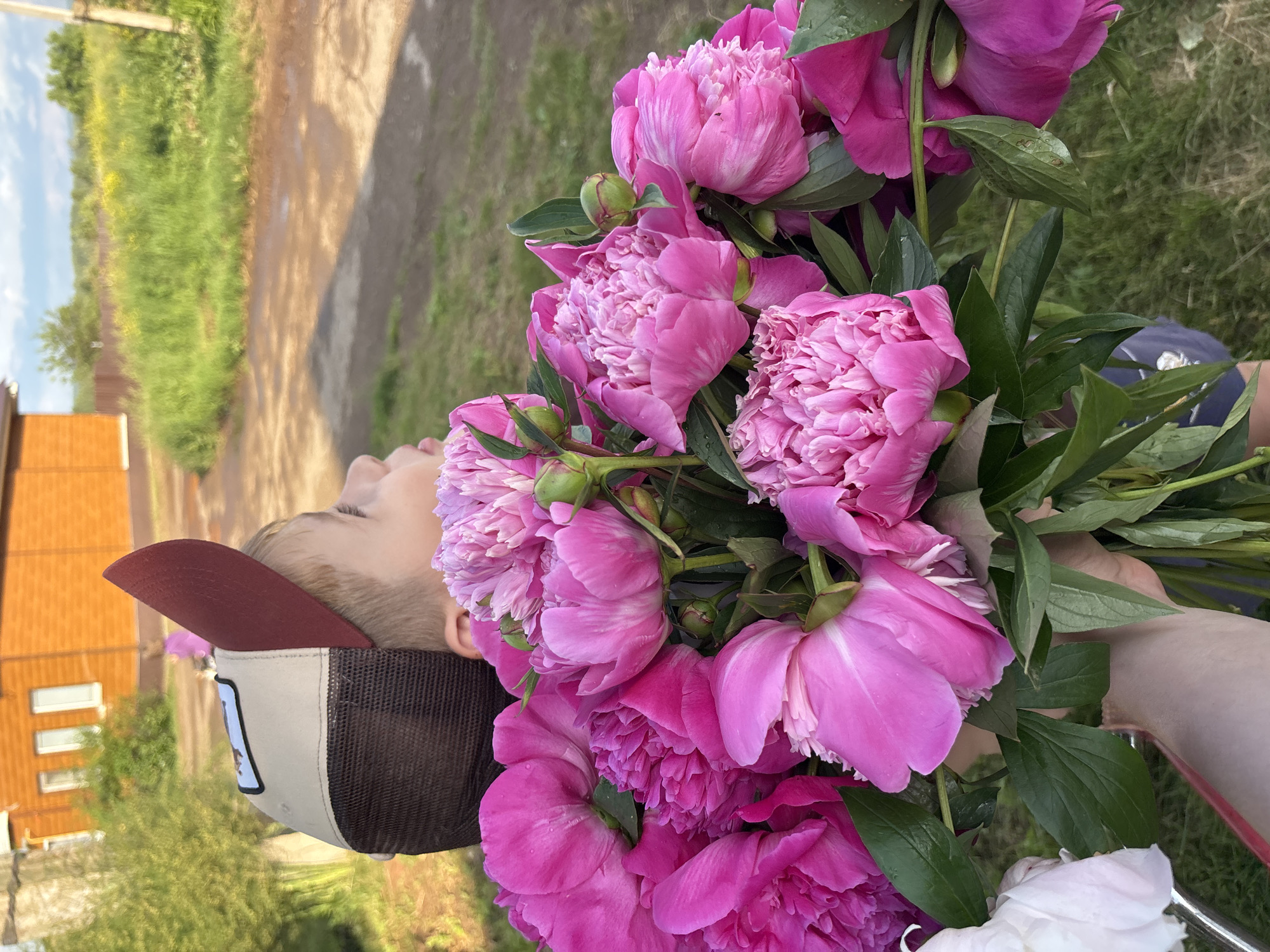 A young boy, wearing a beige and maroon baseball cap, is holding a large bouquet of vibrant pink peonies. He is looking upwards and to the right. In the background, a wooden house with visible windows is situated on the left side of the frame. The scene appears to be outdoors, with green grass and a dirt path visible. The boy and the flowers are the primary subjects. The location is identified as the city of Niva, Russia.
