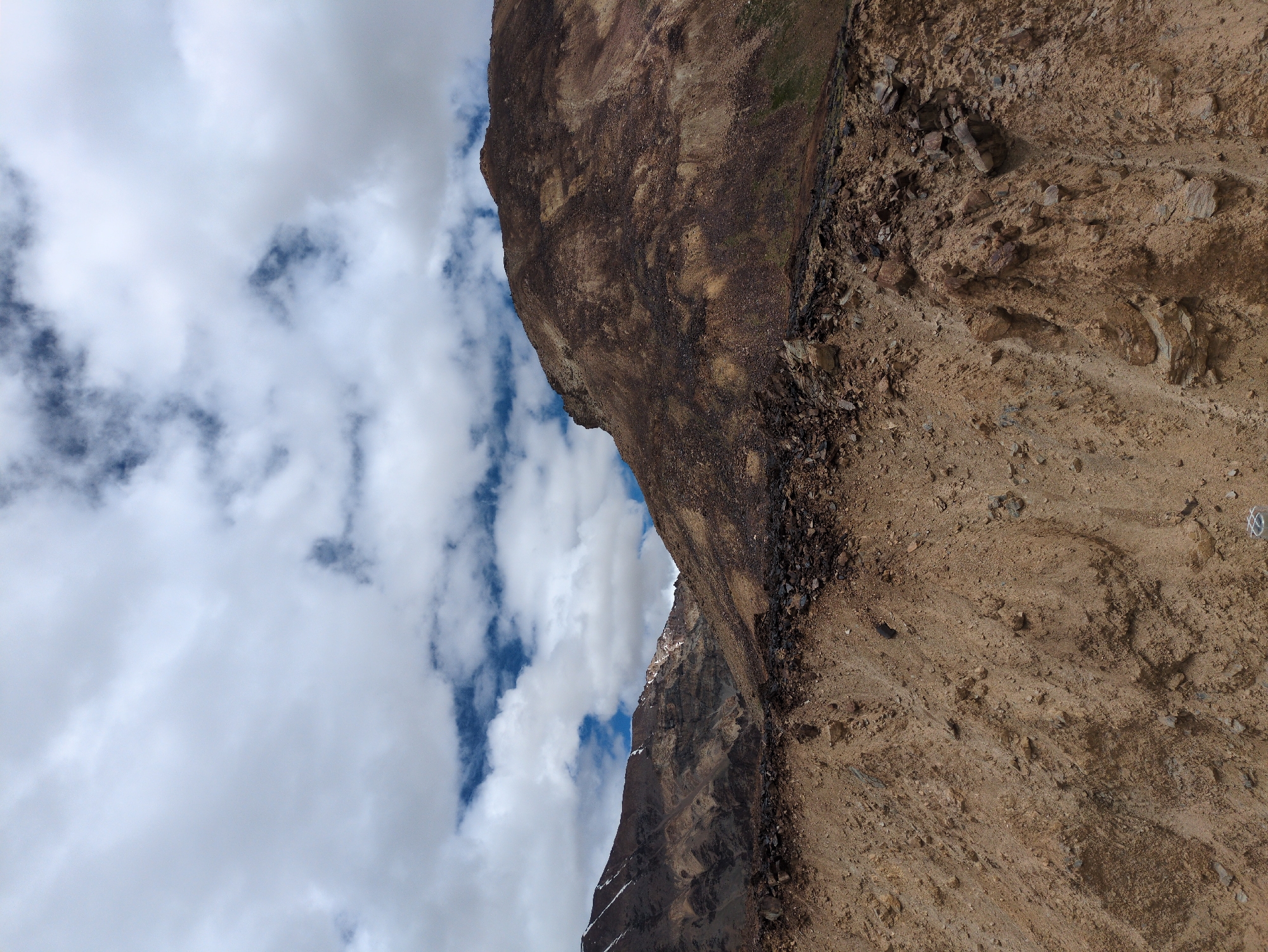 The image shows a mountainous landscape with a cloudy sky. In the foreground, there is a steep, rocky slope with brown soil and scattered stones. The midground features more rugged, brown-colored mountains with visible layers of rock and some patches of snow near the peaks. The background is dominated by a sky filled with dramatic white and gray clouds, with patches of blue sky visible in between. There are no people or man-made structures visible in the image. The scene appears to be in a natural, arid, or semi-arid mountainous region. The location is identified as Killingsarai, India.