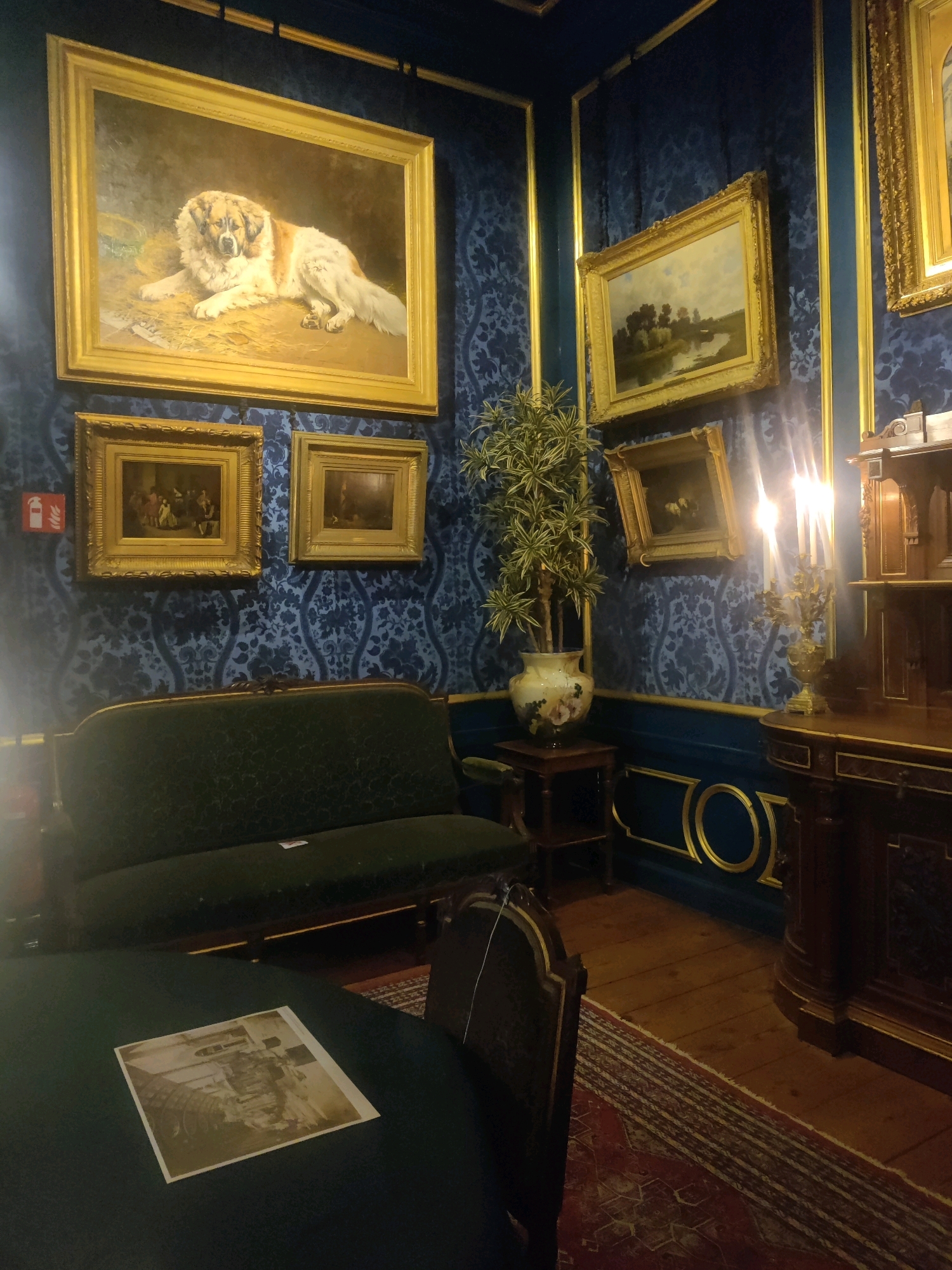 The image depicts an opulent room, likely a museum or a well-preserved historical interior, featuring richly patterned blue wallpaper with gold accents. Dominating the wall is a large framed painting of a fluffy dog, possibly a Bernese Mountain Dog, lying on straw. Below this, two smaller framed paintings hang side-by-side. To the right, another framed landscape painting is displayed.
A dark green velvet sofa is positioned against the wallpapered wall. In the foreground, a round table with a dark surface holds a framed photograph or print. A wooden chair with gold trim is partially visible. To the right, a wooden cabinet or dresser stands prominently, adorned with ornate carvings and topped with candles in a candelabra. A potted plant in a decorative vase sits on a small wooden stand. The floor is made of polished wooden planks, and a patterned rug is laid down. The overall impression is one of refined elegance and historical significance.
This specific room is located in the Museum Van Loon in Amsterdam, Netherlands. It is a historic canal house museum that showcases the lifestyle of a wealthy Amsterdam merchant family.
