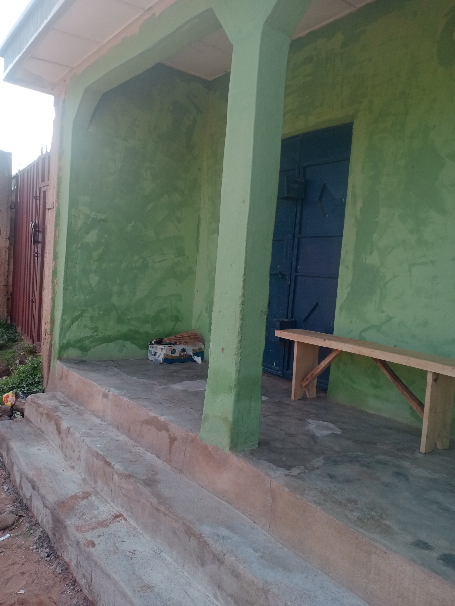 The image depicts the entrance to a building with green-painted walls and a light-colored ceiling. A large blue door is visible on the right side of the scene. In front of the door, there is a simple wooden bench. To the left of the entrance, a brown metal gate is present, with some overgrown vegetation in front of it. Steps lead up to the entrance, and the ground appears to be made of concrete and dirt. A cardboard box containing brooms and other items sits near the left wall. The overall scene suggests an outdoor area, possibly a residential or commercial property in Jalingo, Nigeria.