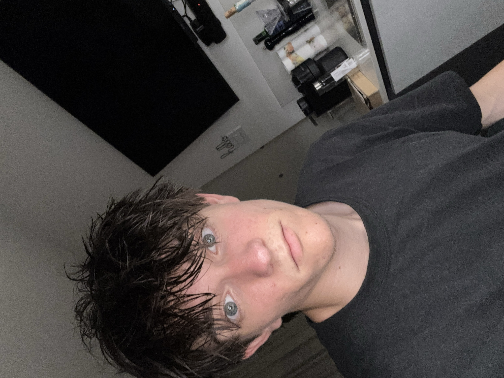 A young person with messy, dark hair and blue eyes is pictured in a close-up, self-taken photograph. They are wearing a dark t-shirt and appear to be indoors, with a black television screen visible in the background, along with some shelving that holds various items including bottles and what appear to be coffee pods. The lighting suggests it is either evening or nighttime. The image does not provide any information that would indicate a specific location within the city of Cupra Marittima, Italy.