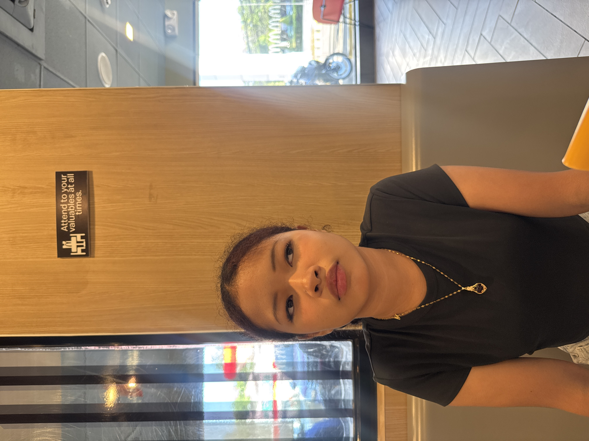 A young woman with dark hair and a neutral expression is seated indoors, looking directly at the camera. She is wearing a black t-shirt and a delicate gold-colored necklace with a small, dark pendant.
The setting appears to be a casual dining establishment, possibly in Davao City, Philippines. Behind the woman, there is a light-colored wooden partition. Affixed to this partition is a black sign with white text that reads, "Attend to your valuables at all times," along with a small, stylized logo above it.
Beyond the partition, a large window reveals an outdoor scene with bright daylight, showing green foliage, suggestive of trees or bushes, and faint glimpses of what appears to be a red bag and parts of a bicycle or scooter. Inside, above the wooden paneling, a gray-tiled wall with a geometric pattern and some light fixtures are visible. To the woman's right, a portion of a yellow or orange cup is seen. The bright natural light streaming in suggests it is daytime. The woman's posture is relaxed, and her expression is calm.