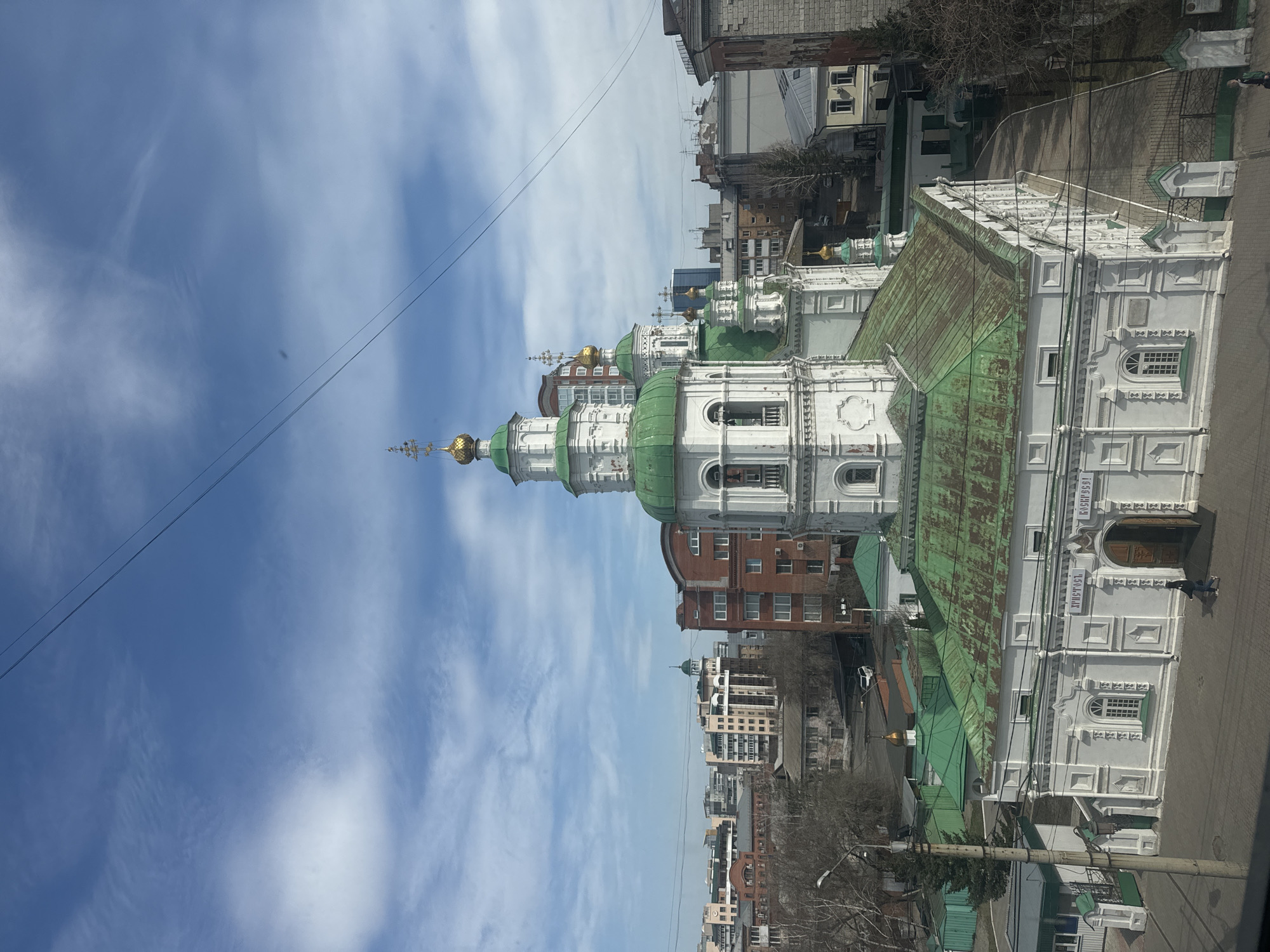 Here is a detailed description of the image:
The image shows an elevated, outdoor view of the Intercession Cathedral in Krasnoyarsk, Russia, set against a partly cloudy sky. The cathedral's architecture stands out, featuring white walls with green roofs and domes topped with golden crosses. Several building are visible in the background, ranging from brick structures to modern high-rise buildings. The urban environment is evident with bare trees interspersed among the structures. A power line runs across the top of the image. A man stands in the foreground.