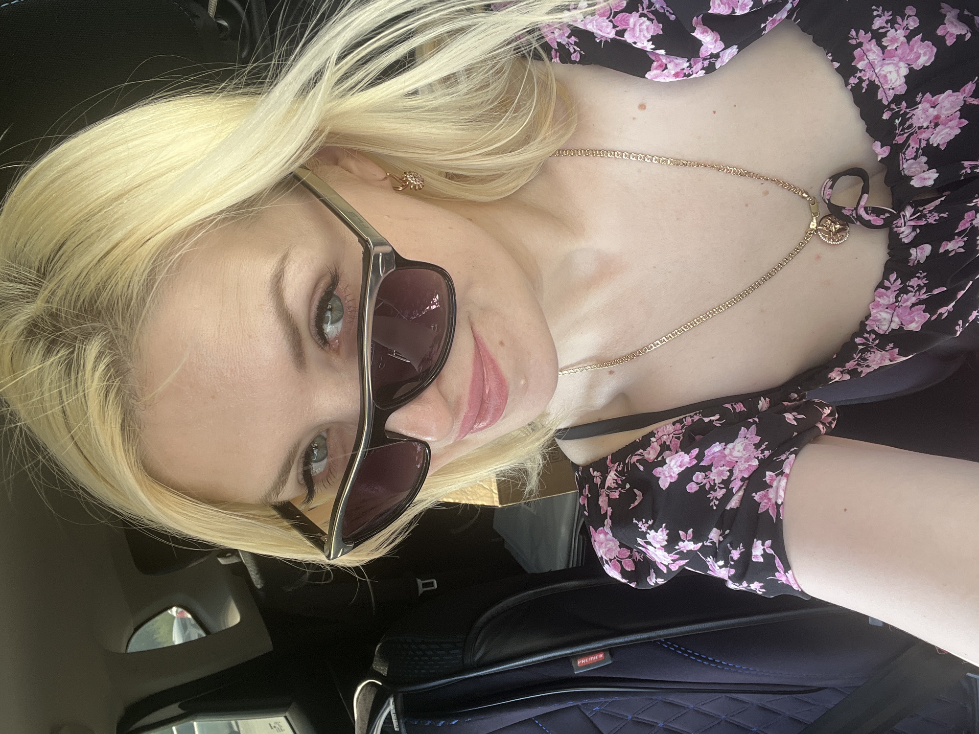A blonde woman wearing sunglasses and a floral-print top is sitting in the passenger seat of a car. Her hair is styled down, and she is looking towards the camera. The background shows the interior of the car, including a seatbelt and a car seat cover. The overall scene suggests a casual outing or travel. There is no information in the image that can be used to determine the location in the city of Tyumen, Russia.
