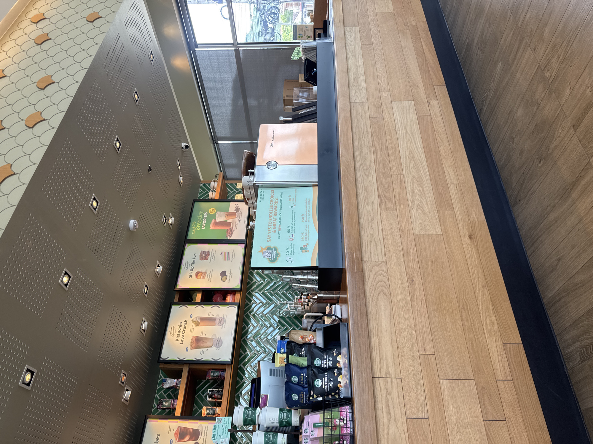 The image shows the interior of a Starbucks coffee shop. A long wooden counter is in the foreground, with various Starbucks products and equipment on top. Behind the counter, there are shelves displaying more products and promotional posters. The ceiling is a metallic grid with recessed lights. The environment appears clean and well-lit, suggesting a typical retail setting. There are no people visible in the image.
