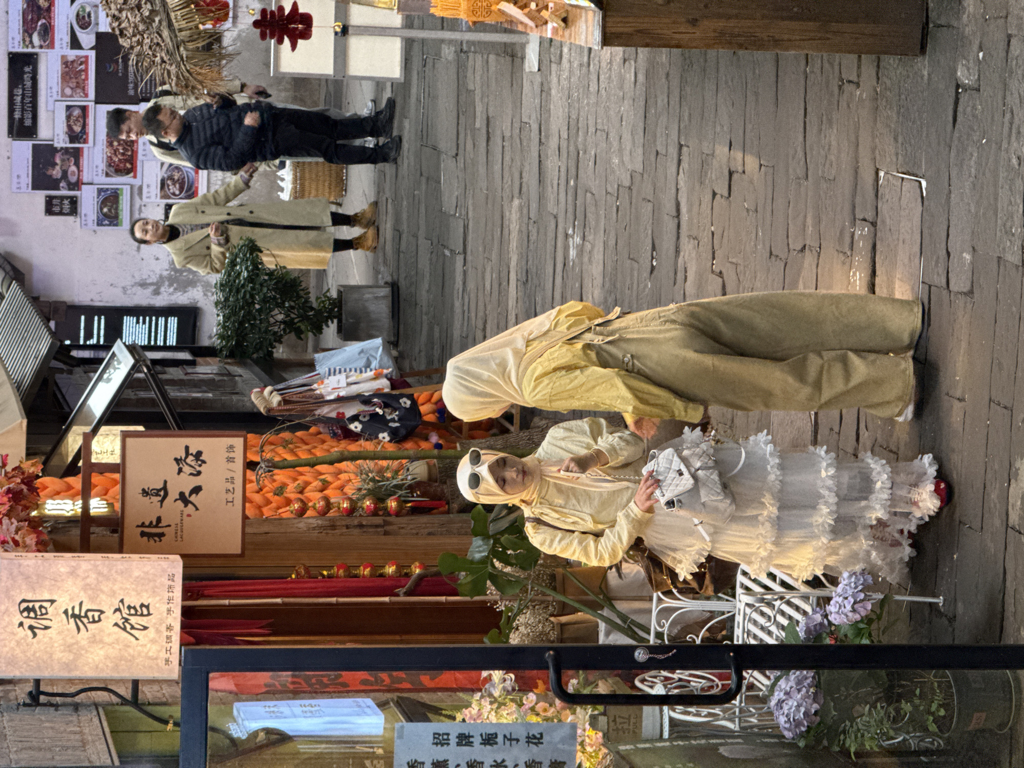 The image depicts a narrow outdoor pedestrian thoroughfare within Yuzhong District, China, characterized by stone-paved ground and buildings housing shops.
In the foreground, two women are positioned mid-conversation. The woman on the left, an East Asian individual, wears a light yellow hijab and a matching long-sleeved top over a tiered white ruffled skirt, complemented by red shoes. She has sunglasses perched atop her hijab and carries a small white quilted handbag with a silver chain strap in her left hand, which rests on the bag. Her right hand is slightly raised. She faces the woman on the right, also an East Asian individual, who wears a light yellow hijab and a beige or light brown jumpsuit. This second woman's back is partially turned to the viewer as she faces her companion.
The immediate background to the left features a shop entrance with a black framed glass door, to the left of which a white ornate metal bench holds potted plants, including purple hydrangeas. Above this entrance, a vertically oriented rectangular white sign bears Chinese characters "调香馆" (Perfume House) and "手工调香 手作饰品" (Hand-blended perfume, handmade ornaments). Red fabric drapes are visible within the shop's entry. Adjacent to this, another shop displays a brown rectangular sign reading "非遗 大漆" (Intangible Cultural Heritage Lacquerware), with "CHINESE LACQUERWARE" in English, and "工艺品 / 首饰" (Crafts / Jewelry). Behind this sign, a decorative installation of vertical orange woven tubes is visible, with various bags hanging from a nearby rack.
Further in the background, a light-colored building wall is adorned with multiple framed posters showcasing food dishes and Chinese text. Three additional individuals are present: an East Asian woman in a long, light beige trench coat, dark pants, and brown boots, standing near a woven basket; and two East Asian men, one in a dark blue puffer jacket looking down at a mobile device, and another behind him in a light-colored shirt or jacket. These individuals appear to be walking or standing casually. To the far right, a partial view of a wooden display stand with a red Chinese knot decoration hanging above it is visible. The ground throughout is paved with irregular dark grey stone slabs.