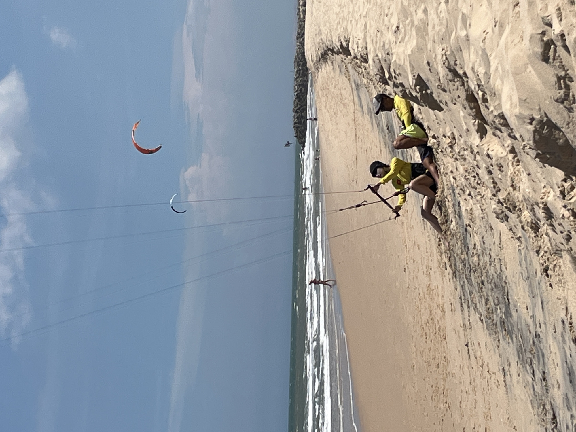 Two individuals are sitting on a sandy beach. One person, wearing a yellow shirt and black shorts, is leaning back on their arms with their legs extended. The other person, also in a yellow shirt and dark shorts, is sitting upright, holding onto a kite control bar. Both are wearing helmets. In the background, there are waves breaking on the shore, and several kites are visible in the sky, including a red kite and a smaller white kite. A person is walking along the water's edge in the distance. A rocky jetty extends into the water. The scene appears to be a beach in Phu Thuy Ward, Vietnam, a location known for kitesurfing. The sky is blue with some scattered clouds.