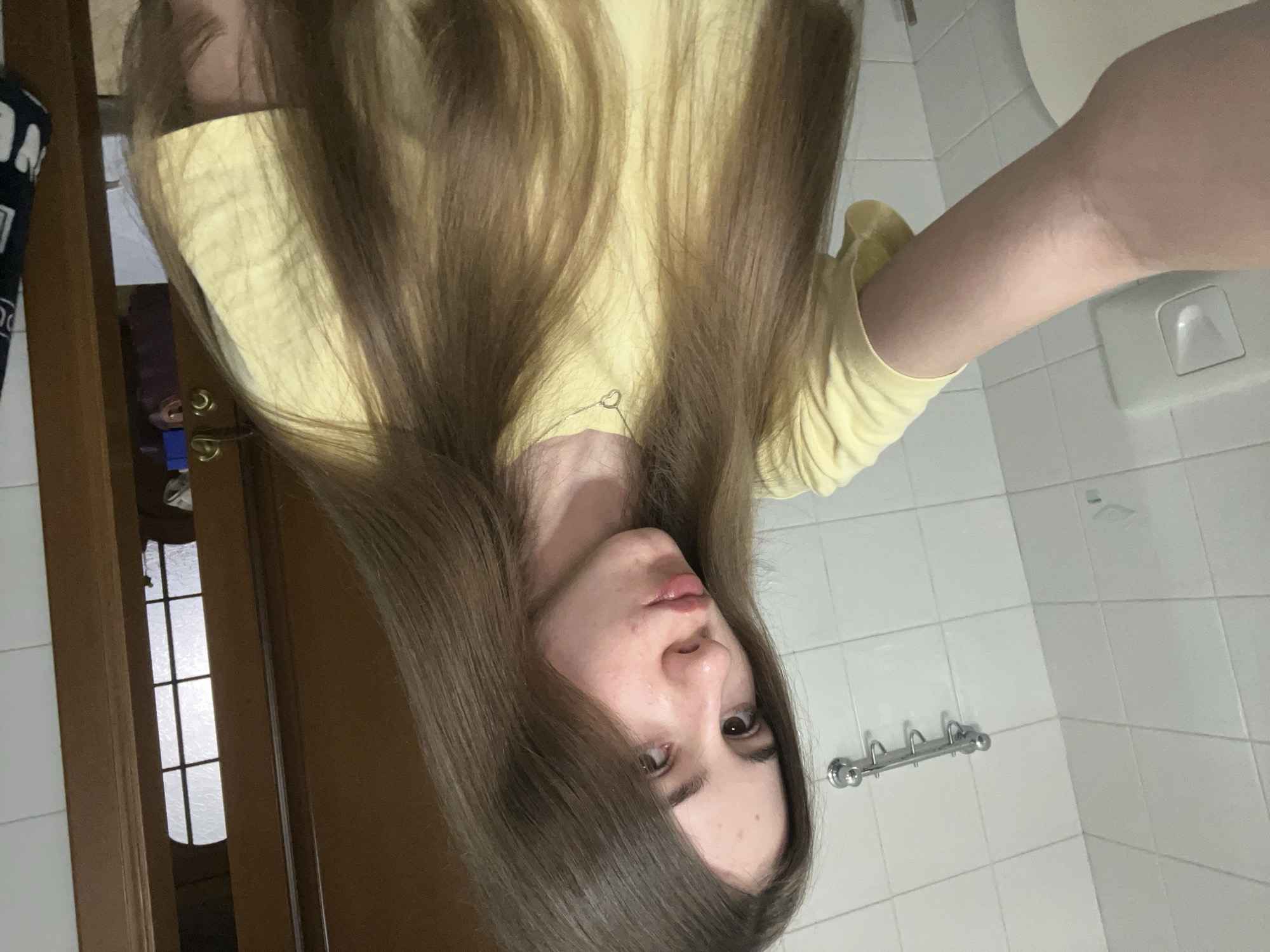 The media file is a portrait-oriented, slightly low-angle, close-up shot of a young person indoors. The person has long, brown hair that partially obscures their face. They are wearing a yellow t-shirt. Their facial features, including brown eyes and lips, are visible. The background suggests a bathroom setting, with white tiled walls and a silver towel rack. To the left, a wooden door frame is visible, with a glimpse of a window with a grid pattern beyond it. A portion of a toilet is also visible in the background to the right. The lighting appears to be artificial, casting soft shadows. The overall mood is casual and candid, possibly a selfie. No text is visible in the image.