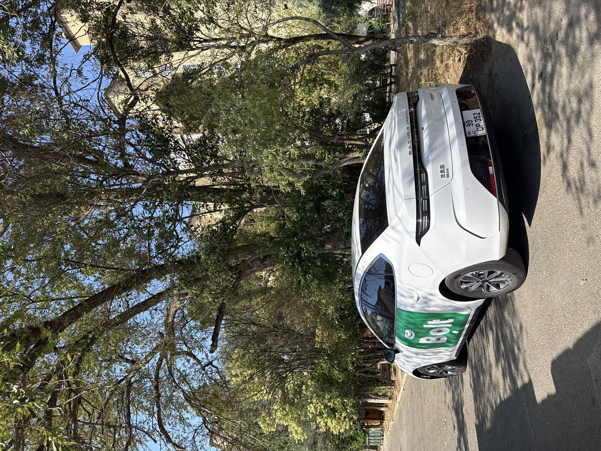 A white sedan with a green "Bolt" logo on its side is parked on a paved street in Baku, Azerbaijan. The car is positioned at a slight angle, facing towards the right side of the frame. The license plate reads "99 QP 352 AZ". Behind the car, lush green trees with dappled sunlight filtering through their leaves create a shaded environment. In the background, a multi-story building with light-colored facade is visible. The scene appears to be a residential area with some benches and foliage on the side of the road. The weather is sunny, and the overall atmosphere is serene.