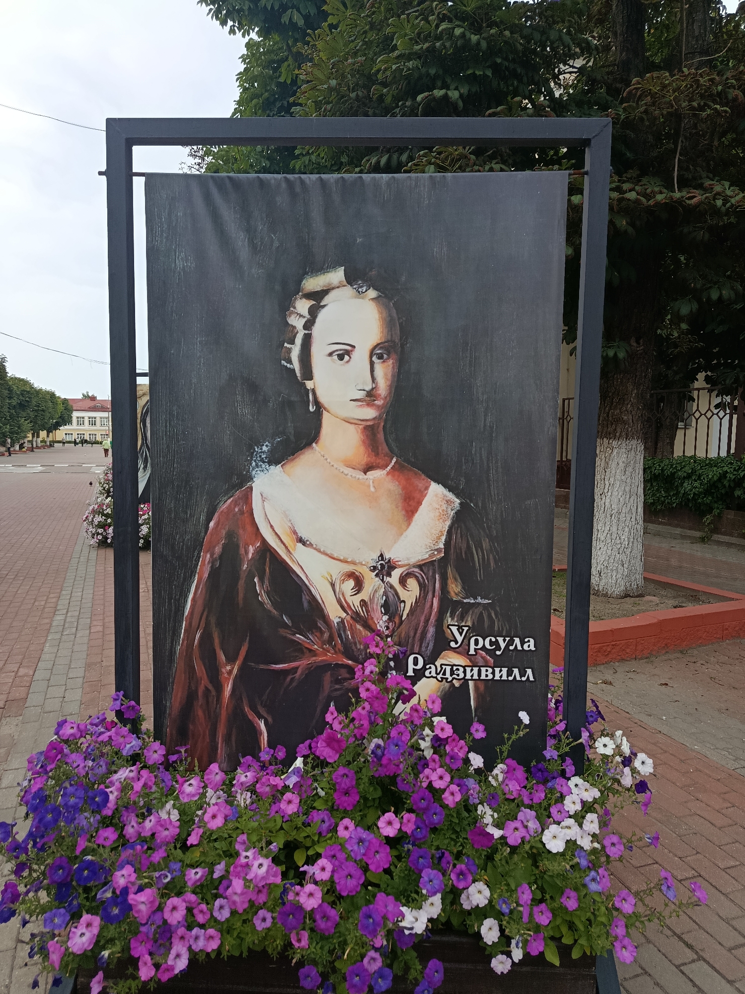 A portrait of a woman in historical attire is displayed on a large banner, framed by a dark metal structure. The banner is placed outdoors, in front of a planter filled with vibrant purple and white flowers. In the background, a street with paving stones is visible, along with buildings and trees. The scene appears to be in the city of Zhlobin, Belarus. The portrait is of Ursula Radziwill, a prominent figure in Belarusian history. The overall impression is one of a public art display, likely in a park or town square.