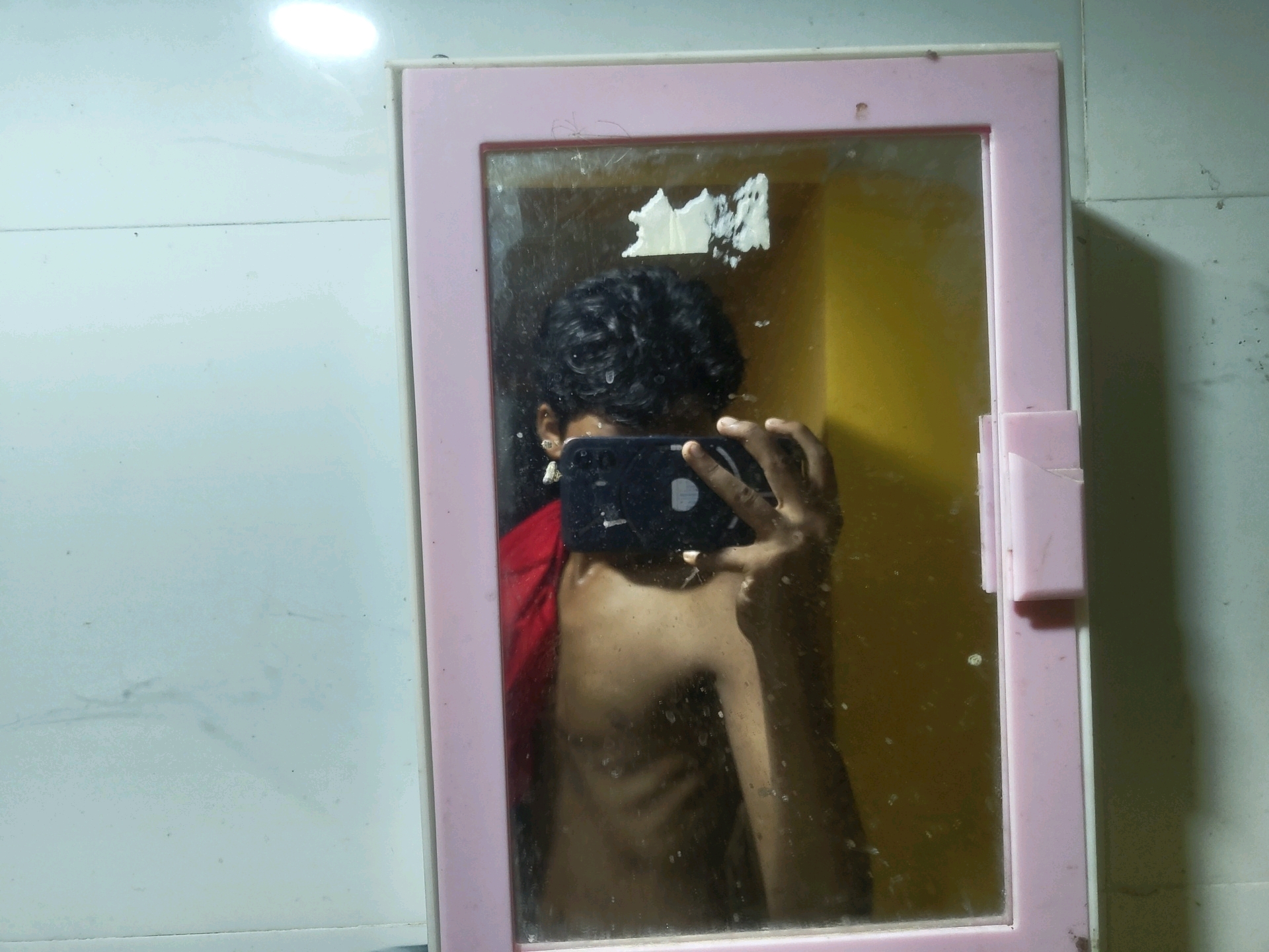 A person takes a mirror selfie in a bathroom. The person is in the center of the frame, holding a phone to take the picture. They are shirtless and wearing a red garment on their left shoulder. The bathroom has a pink medicine cabinet with a mirror. The walls of the bathroom are tiled in white and there is a yellow wall visible behind the person in the mirror. The scene appears to be located in the city of Dahar Buinchha, India.