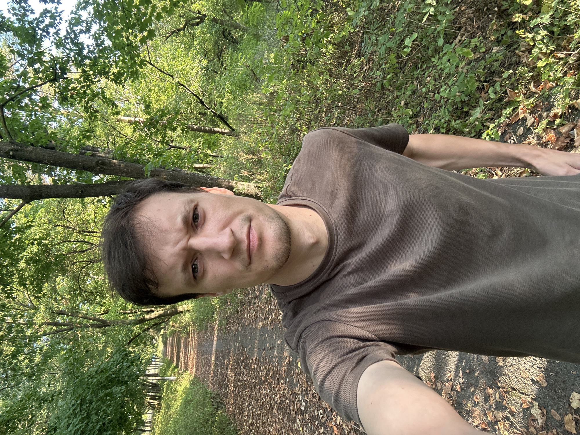 A selfie taken by a man in his early 30s, with brown hair and a short beard, wearing a brown t-shirt. He is standing on a dirt path in a forest, with trees and foliage on either side. The path is covered with fallen leaves. The man is looking directly at the camera with a serious expression. The sun is shining through the trees, casting shadows on the path. The location is a forest path in Makiv, Ukraine.