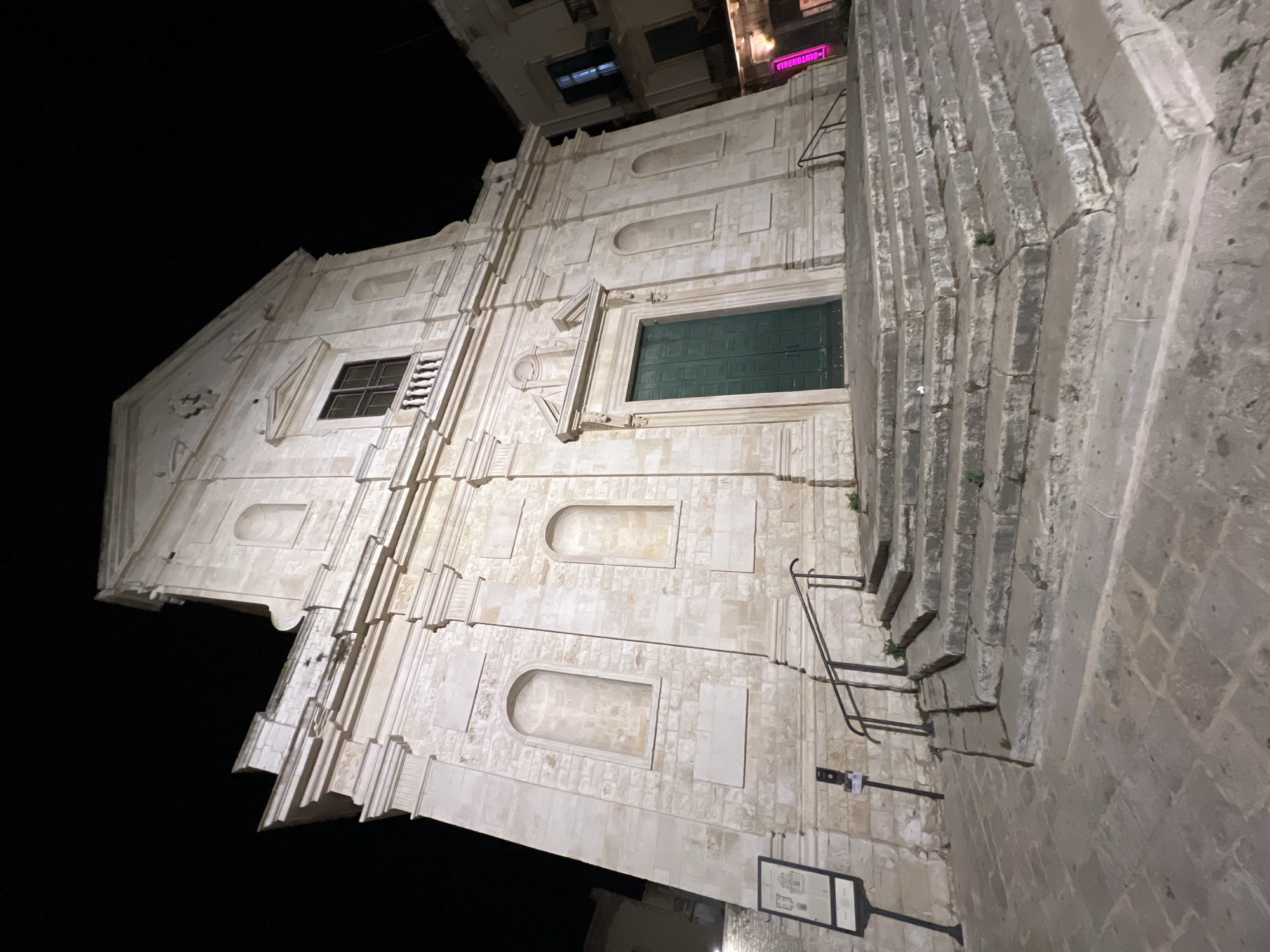 The image shows the exterior of a stone church at night in Bitonto, Italy. The church has a large set of stone stairs leading up to its entrance. The entrance features a green door. Above the door, there is a window with a balcony. The facade of the church is decorated with architectural details, including arched niches and molding. The surrounding area is paved with stone. There are no people visible in the image.