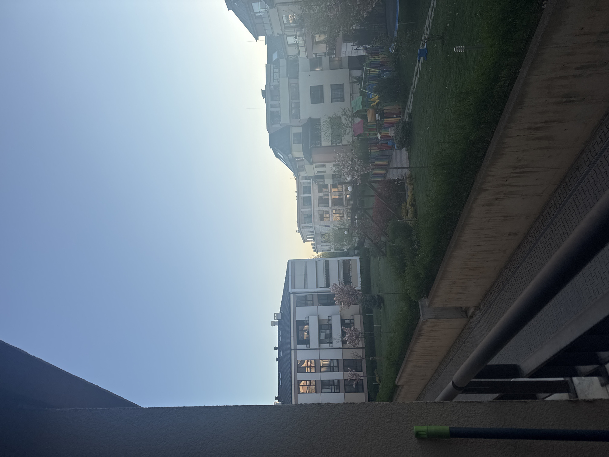 View from a balcony in a residential area of Sofia, Bulgaria, with multi-story buildings facing each other across a green space containing a playground. The sky is light blue and clear. A wall and metal railings are visible in the foreground. A green-tipped pole rests on the wall.