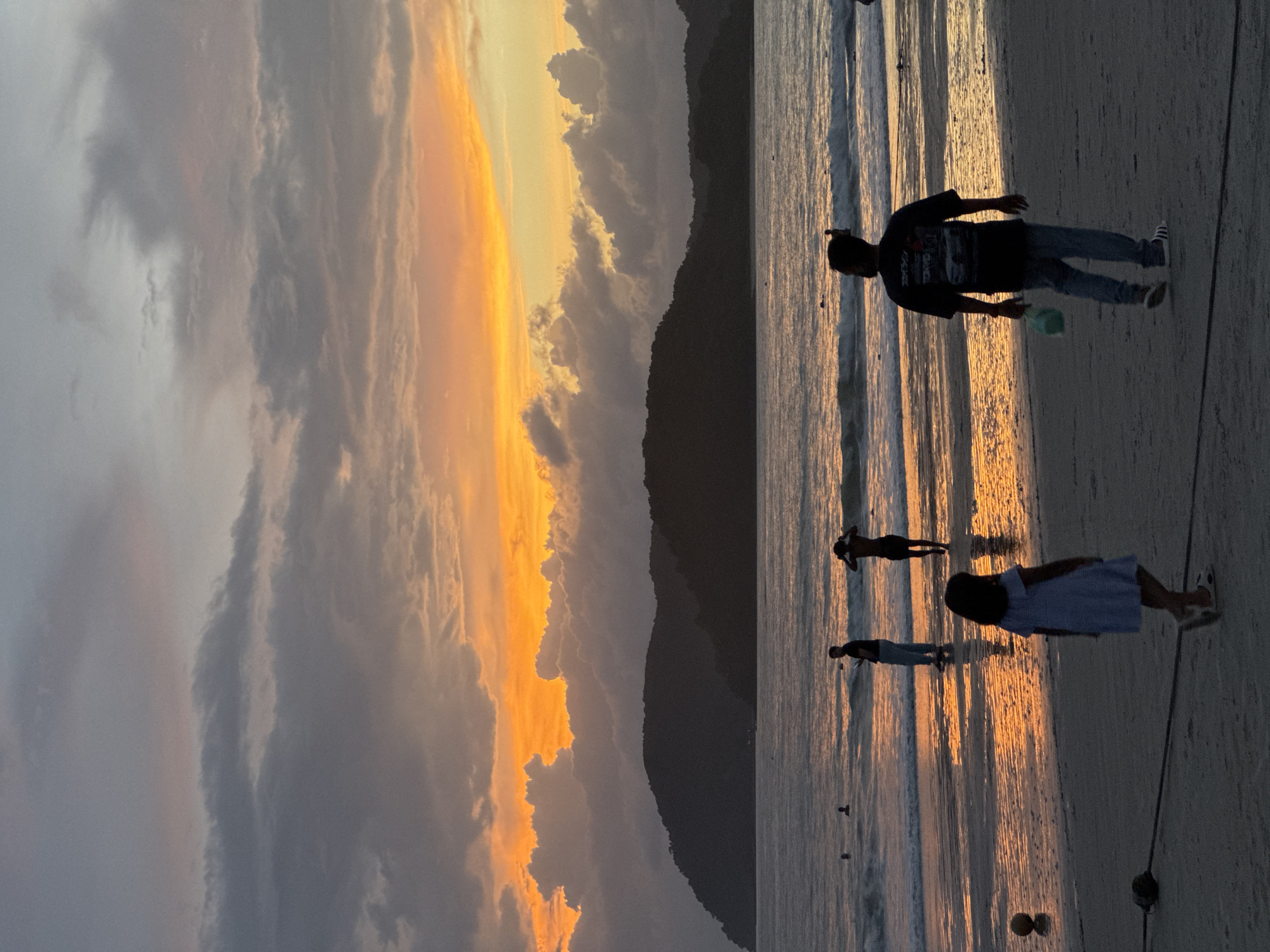 This photograph captures a sunset scene on Cenang Beach, Malaysia. The sky is painted with vibrant hues of orange and yellow, contrasted by dark, silhouetted clouds and the distant, dark outline of a mountain range. The ocean is calm, with gentle waves lapping at the shore, reflecting the fiery colors of the sky.

In the foreground, a young girl in a light blue dress walks away from the camera, her silhouette dark against the wet sand. To her right, a man in a dark t-shirt and jeans walks towards the water, carrying a light green bag. Further out in the water, several people are visible, some swimming and others standing. One man is seen with his hands on his head, facing away from the camera.

The beach itself is sandy, with a few objects visible, including a yellow ball near the water's edge and a dark rope extending across the sand. The overall atmosphere is serene, with the natural beauty of the sunset dominating the scene.