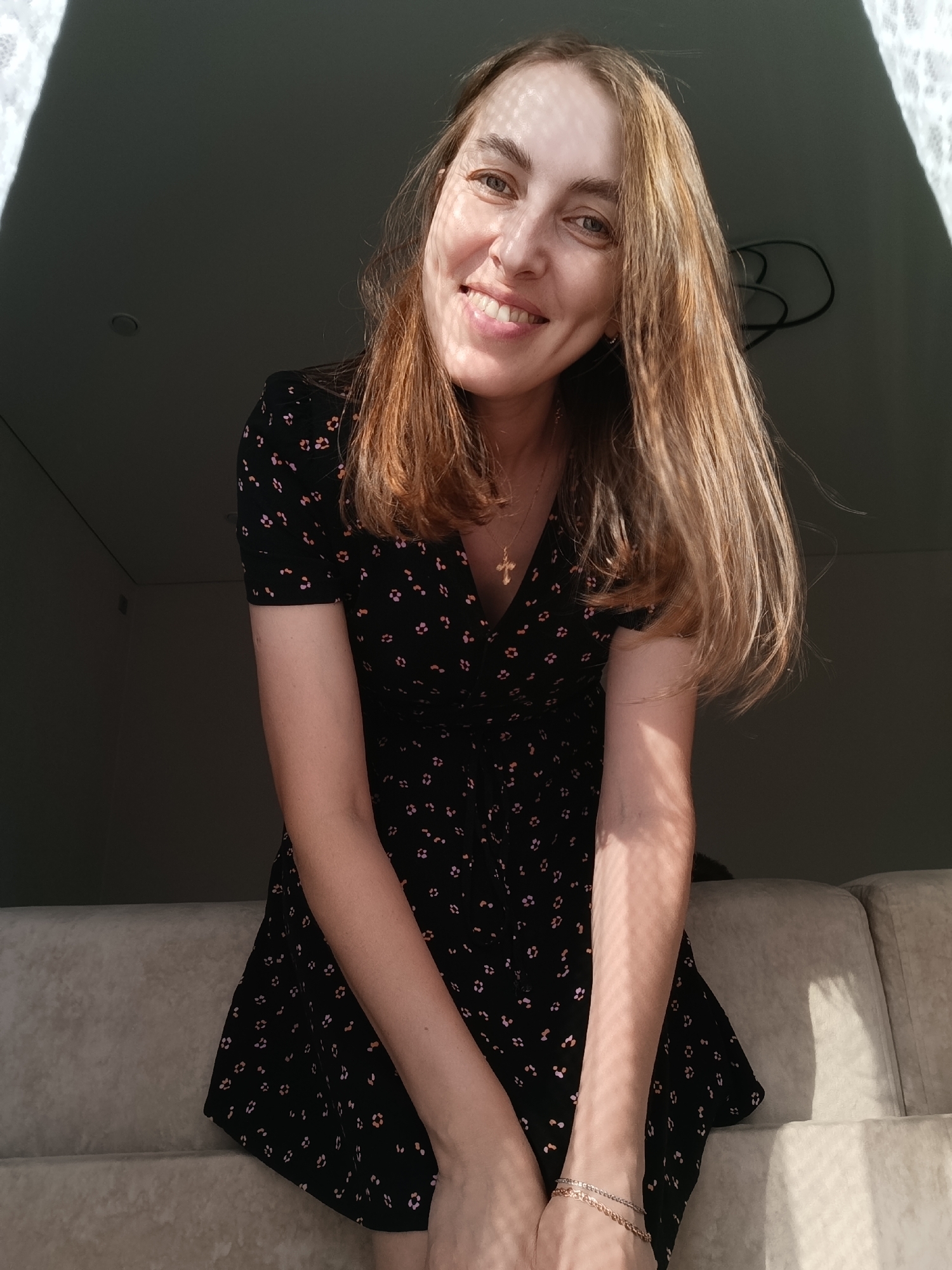 A fair-skinned woman with light brown hair smiles at the camera. She is wearing a short-sleeved black dress with a pattern of small pink flowers. A gold necklace with a cross pendant is visible at her neckline. A gold bracelet is on her left wrist. Light and shadow patterns, likely from a window covering, are cast across her face and arms. The setting is indoors, with a beige sofa visible in the foreground and a dark background behind the subject. A ceiling light is visible in the background. The location is identified as the city of Chaikovsky, Russia.