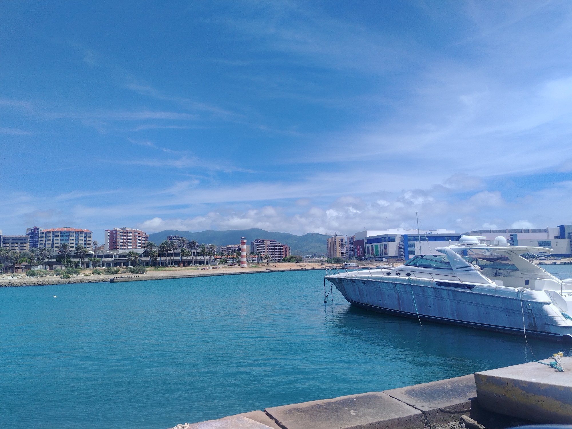A bright, sunny day features a large, white yacht docked in the foreground on the right side of the frame, with its bow pointing towards the left. The yacht appears to be a motor yacht with a sleek, modern design, featuring multiple levels and windows. The water in the harbor is a vibrant turquoise blue. In the mid-ground, a striped lighthouse stands out against a backdrop of buildings and palm trees lining the shore. The buildings appear to be a mix of residential and commercial structures, with some showing a red brick facade and others modern glass and steel construction. Beyond the city skyline, a range of mountains under a partly cloudy sky forms the background. The sky itself is a clear blue with scattered white clouds, some of which are wispy and thin, characteristic of fair weather. The scene suggests a coastal urban environment, likely a harbor or marina in Porlamar, Venezuela, given the architectural styles and lush vegetation. A concrete dock or quay runs along the bottom edge of the frame.