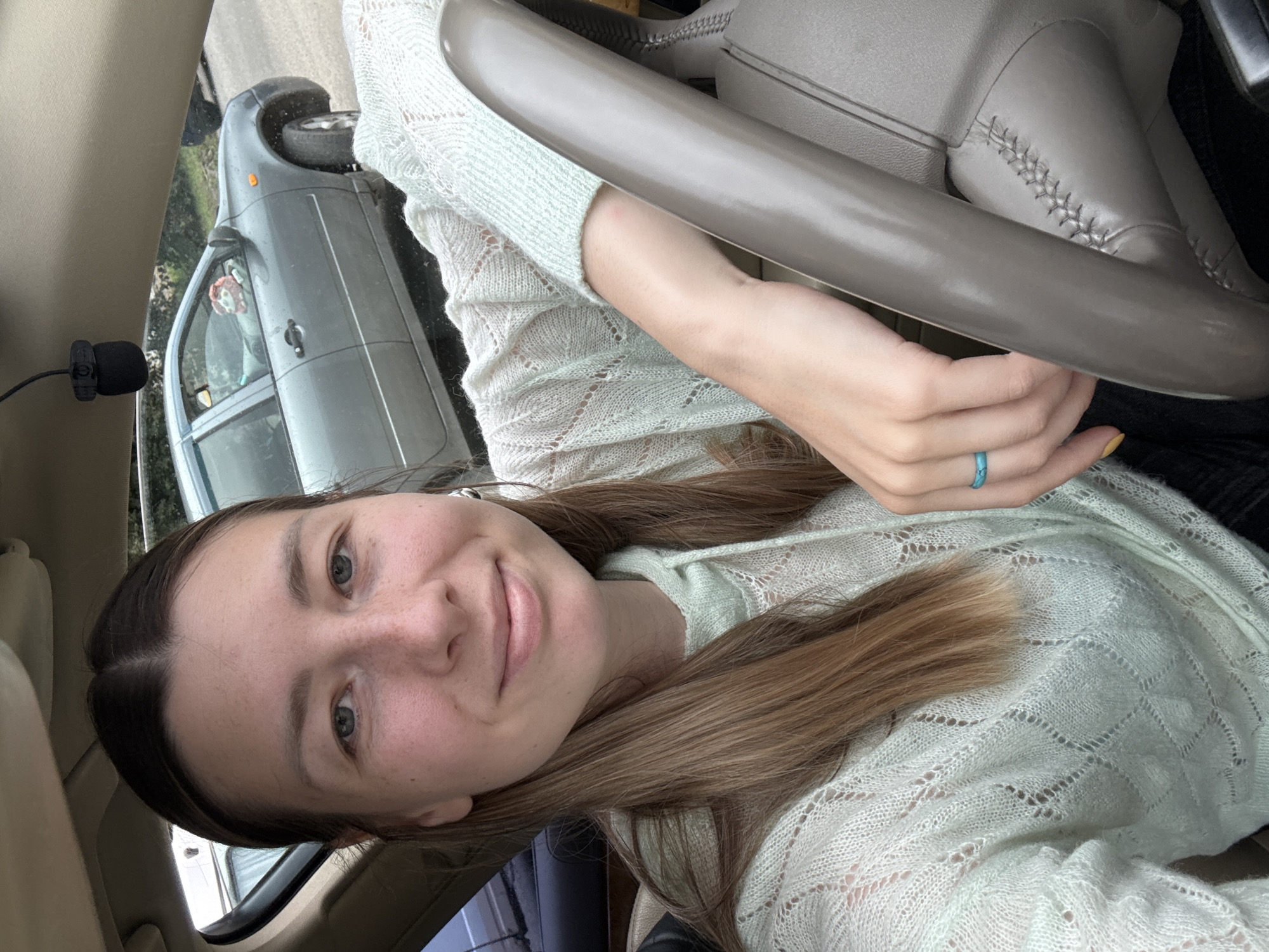 A young woman with brown hair, wearing a light green knitted sweater, is taking a selfie inside a car. She is in the driver's seat, with her left hand resting on the steering wheel. A turquoise ring is visible on her left ring finger. In the background, through the car window, another car is visible on a paved road, suggesting an urban or suburban setting. The overall scene is outdoors.