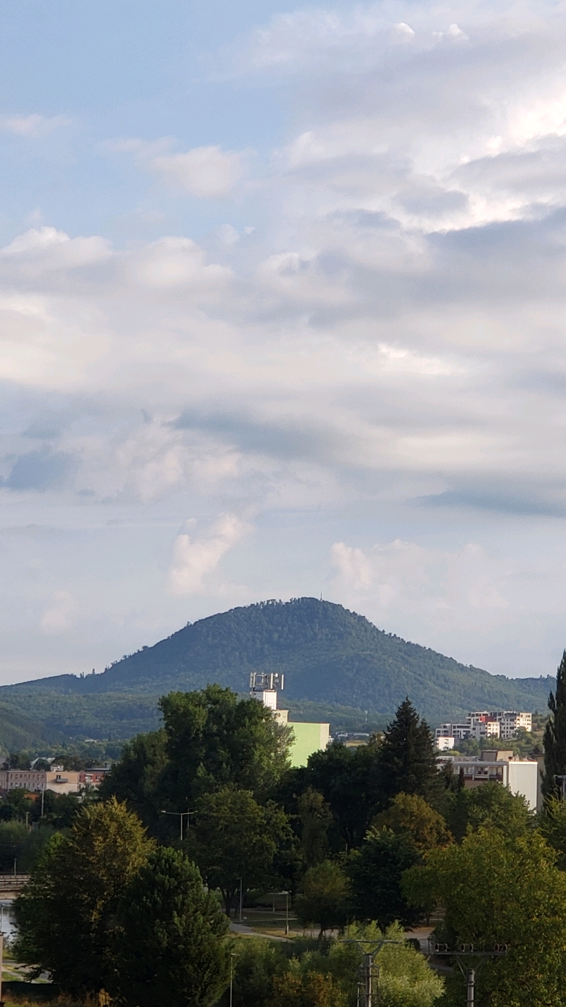 The image is a vertical, eye-level shot of a vast green hill in the background, capped with trees and a radio tower. In the middle ground, a cluster of buildings, including a light green structure topped with communication antennas, is visible. Further down, a dense forest of trees occupies the foreground, with some buildings peeking through. The sky is filled with scattered white clouds against a pale blue backdrop. The scene appears to be a panoramic view of the city of Prešov, Slovakia, with the prominent hill likely being Šariš Castle Hill.