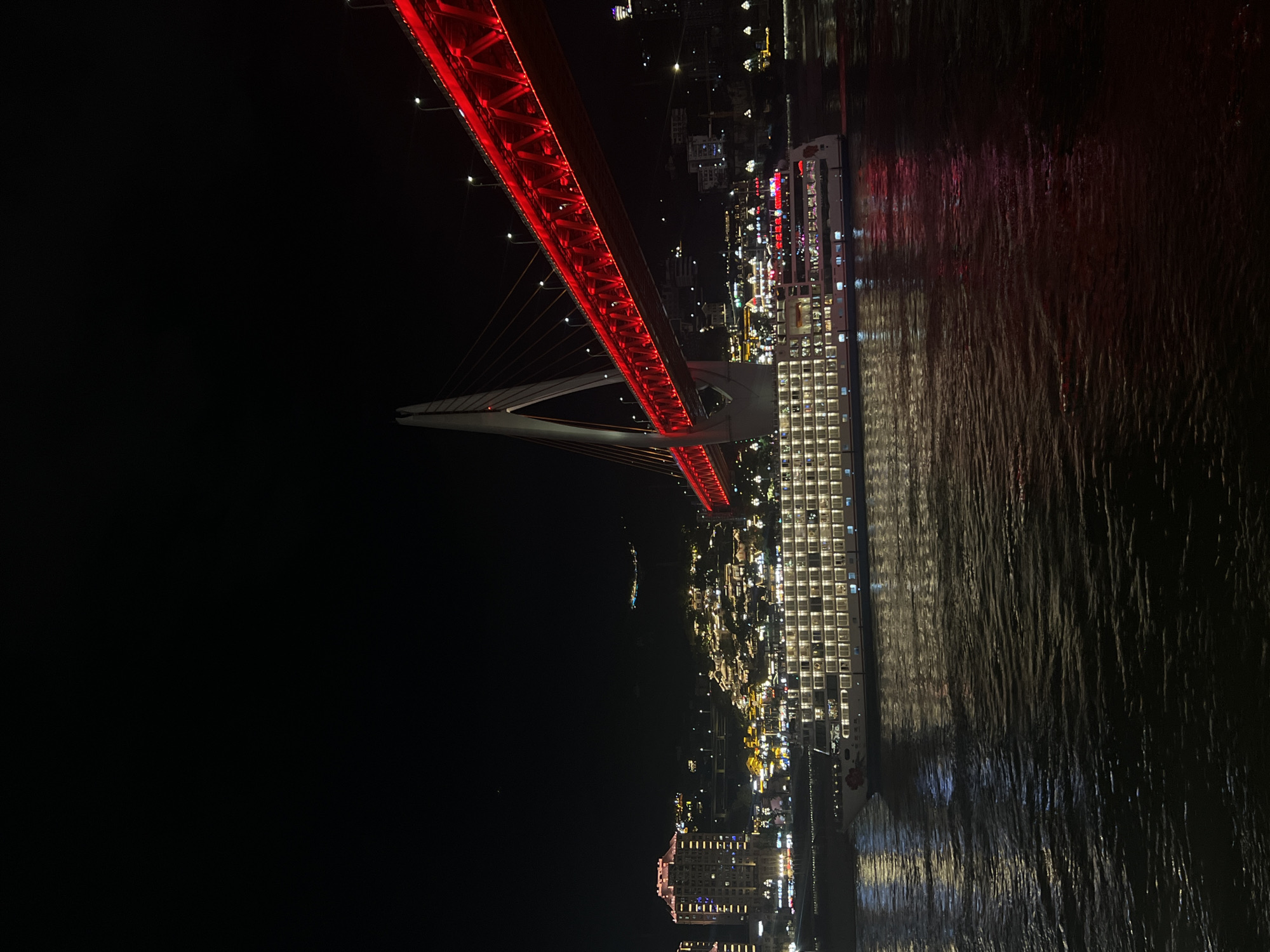 A wide shot captured at night shows the Hongyadong Bridge in Yuzhong District, China, illuminated with red lights. A large, multi-story cruise ship, lit with numerous windows, sails on the dark water in the foreground. The cruise ship has a floral design on its bow. In the background, the cityscape of Yuzhong District is visible, with many buildings and lights cascading down a hillside. The sky is completely dark.